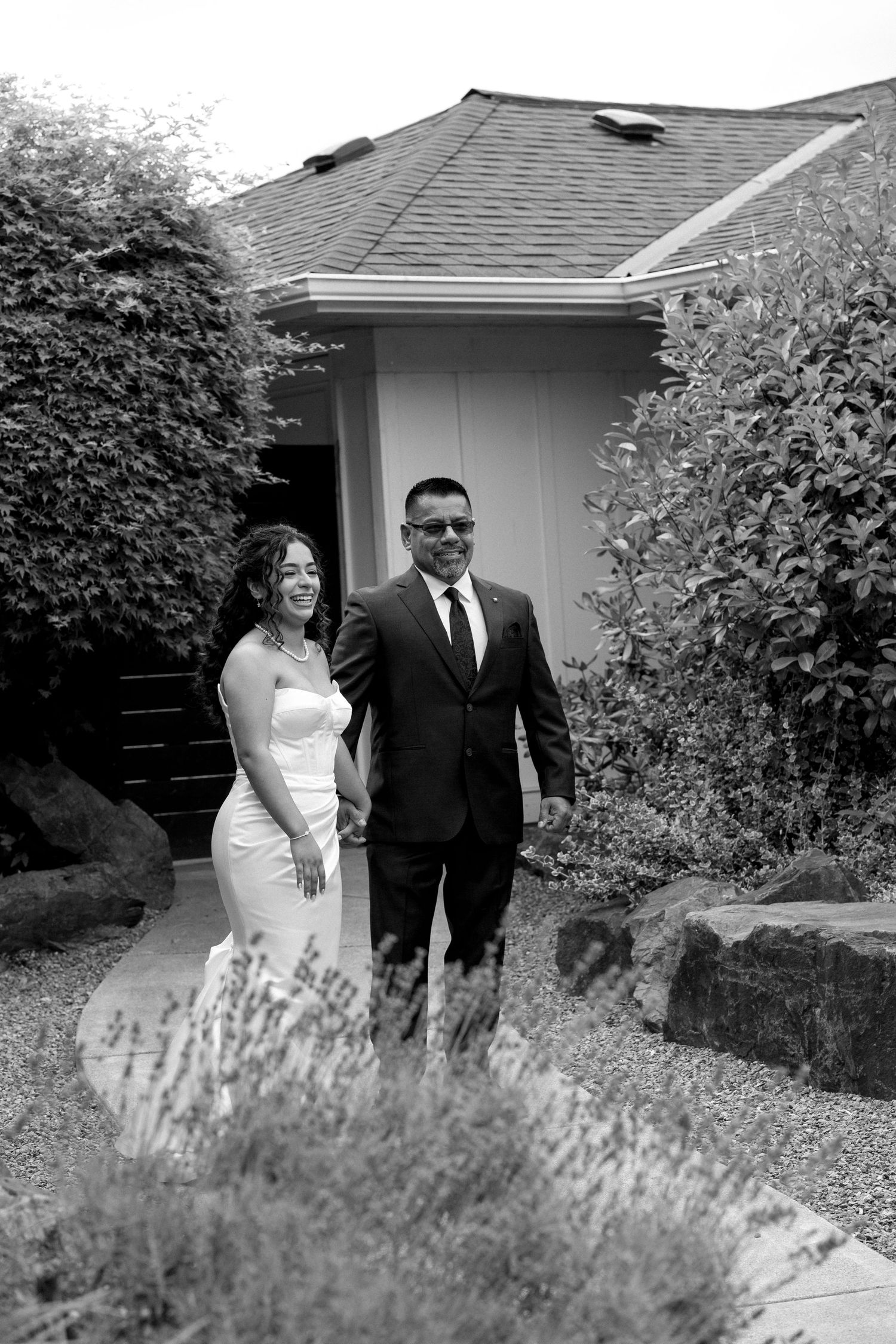 Black and white photo of bride and groom walking hand in hand through garden pathway.
