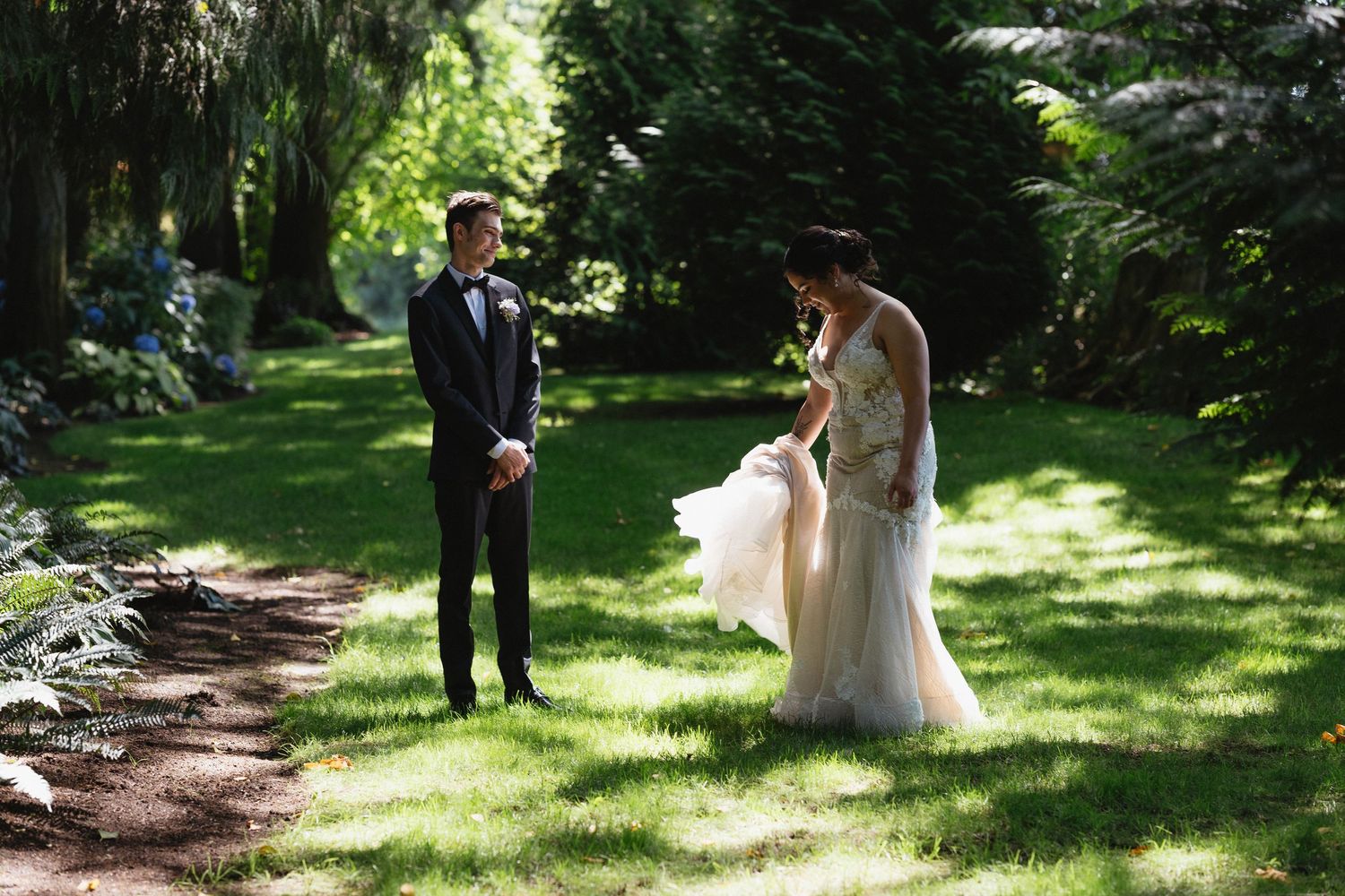 Groom in black tuxedo watches as bride in elegant gown adjusts her dress on a garden path.