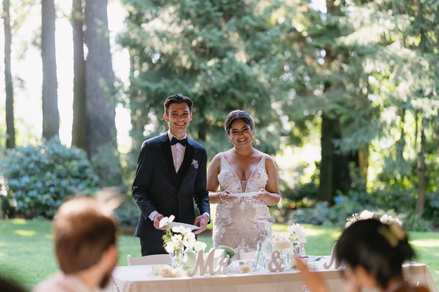 Smiling bride and groom stand together at their outdoor wedding reception table decorated with white flowers.