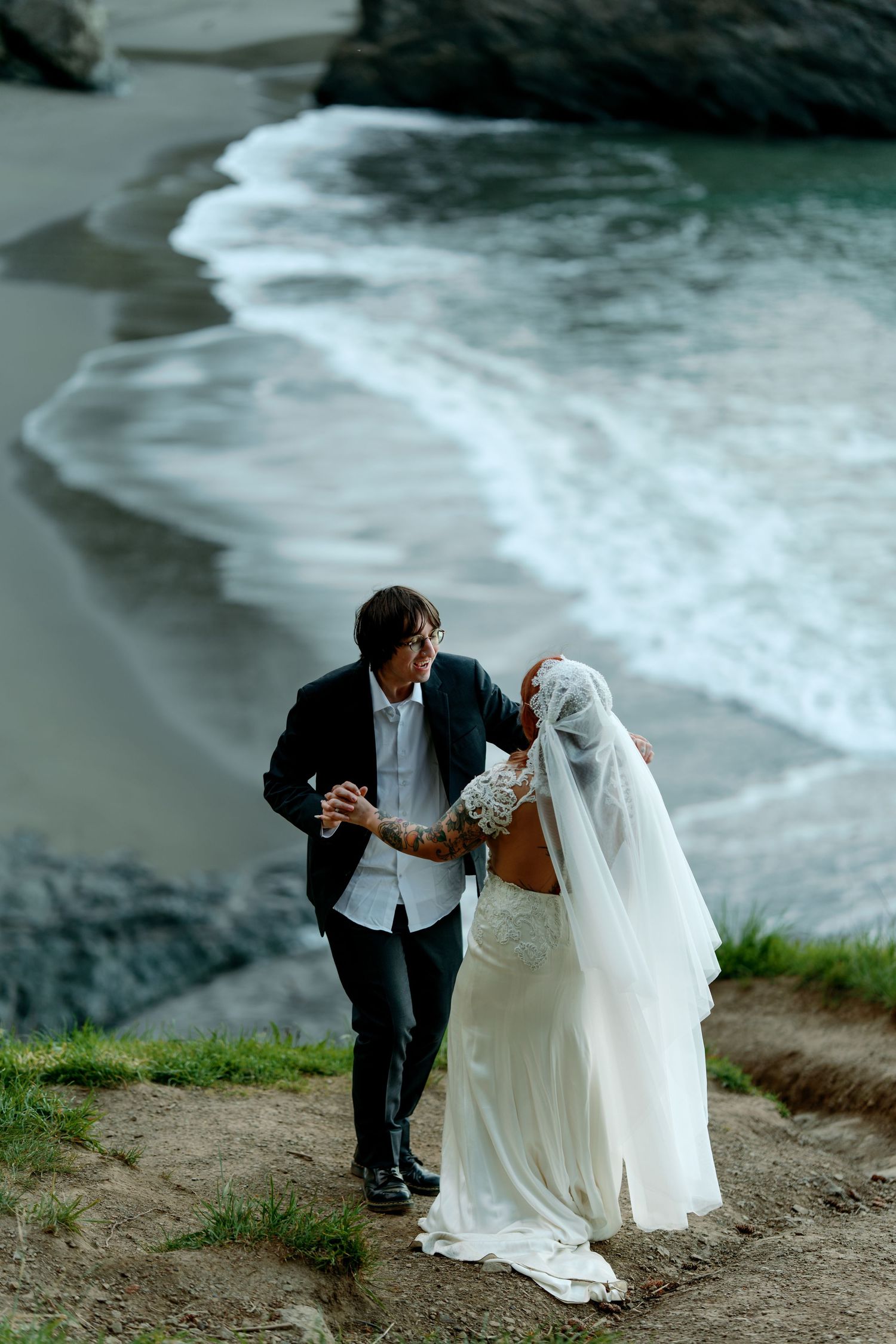 Bride in white lace gown and groom in black suit walk along a cliff overlooking a sandy beach.