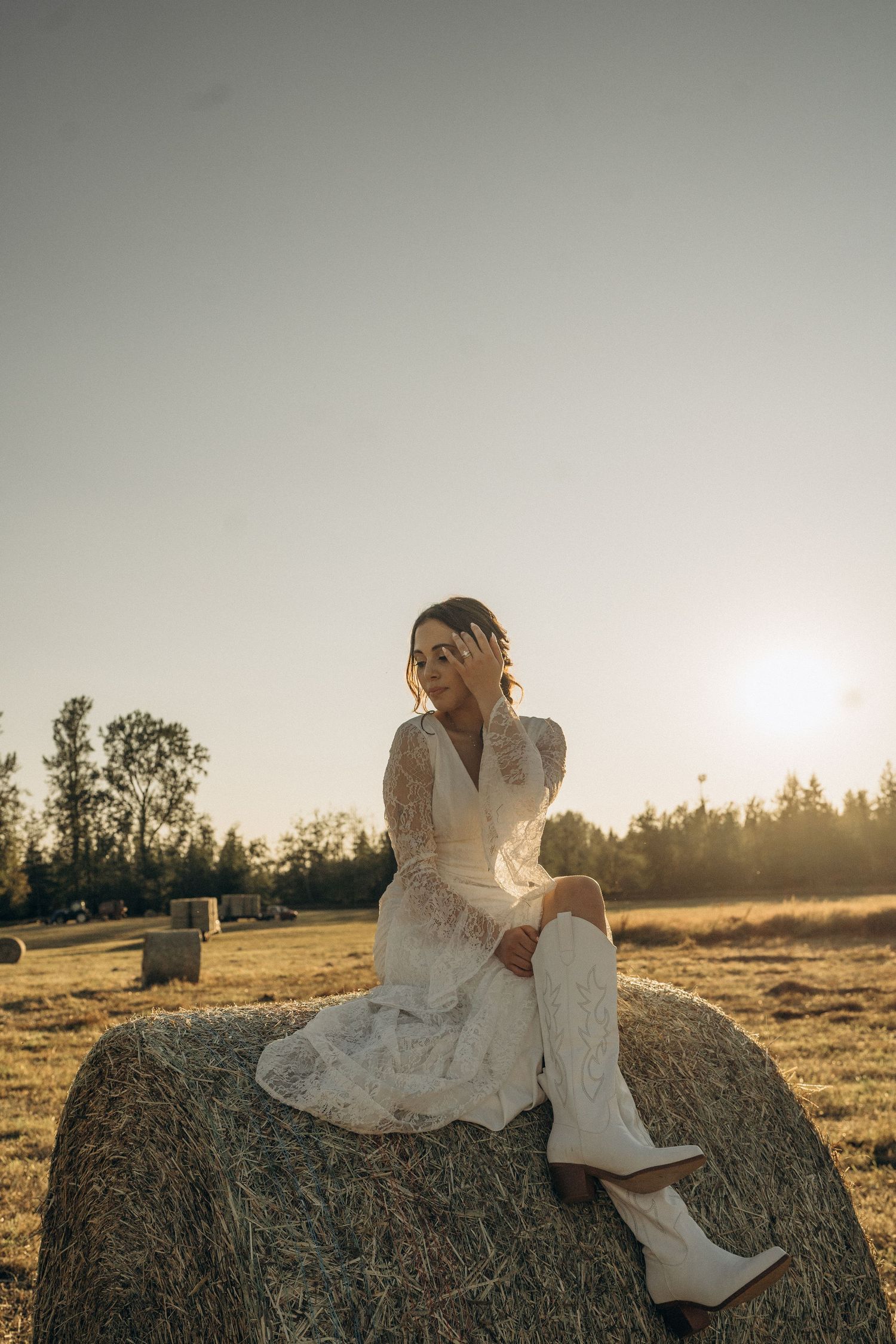 Bride in white lace dress and cowboy boots sits on a hay bale during sunset.