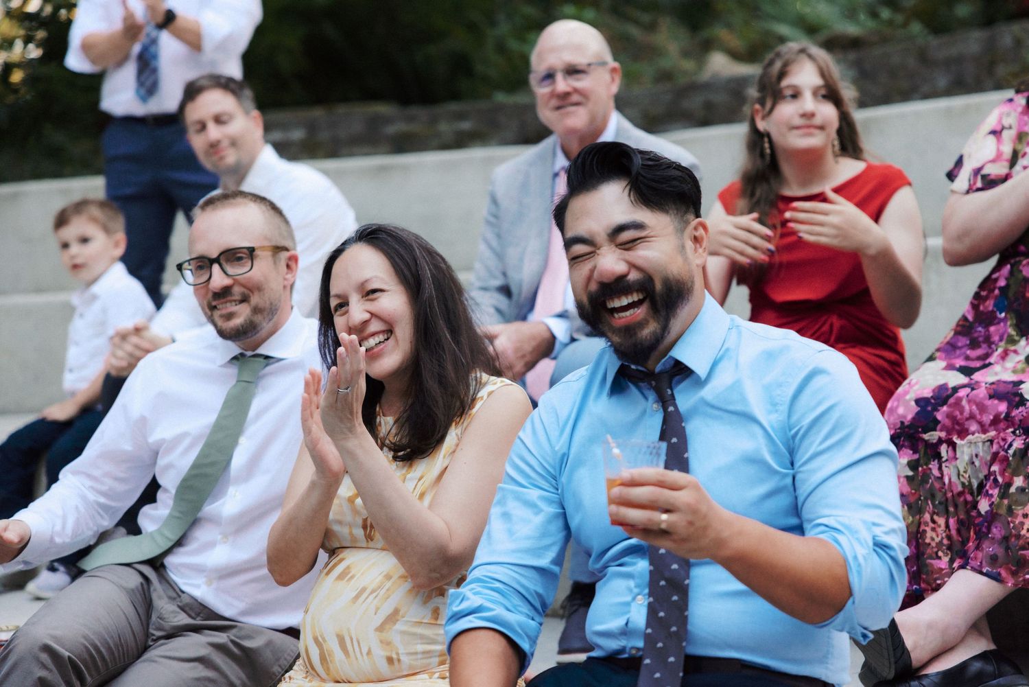 Wedding guests laughing and clapping while seated outdoors during ceremony celebration.