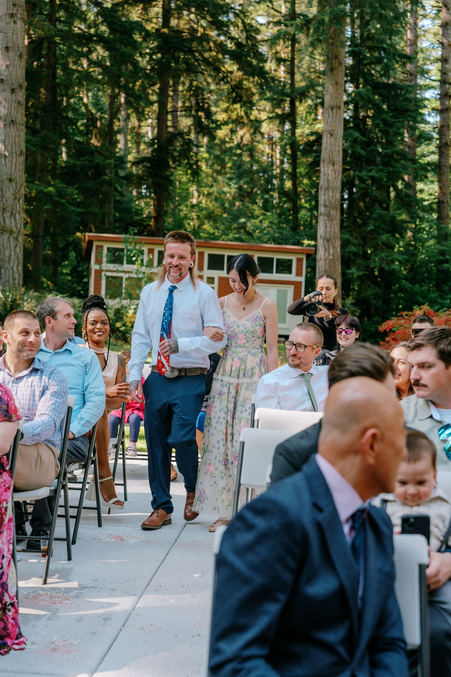 Smiling couple walks down wedding aisle past cheering guests at outdoor forest ceremony.