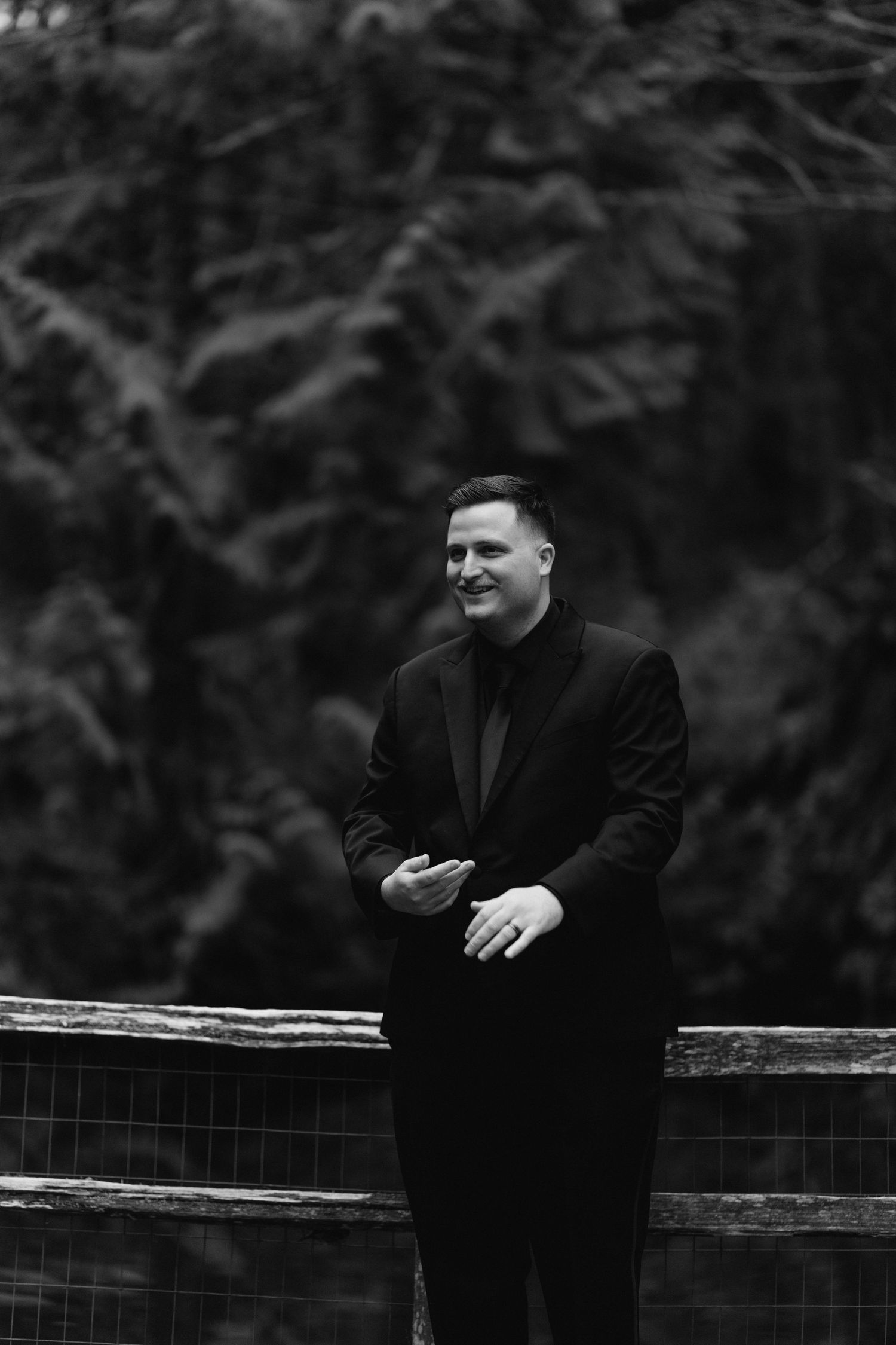 Handsome groom in all-black suit stands confidently by fence in black and white portrait.