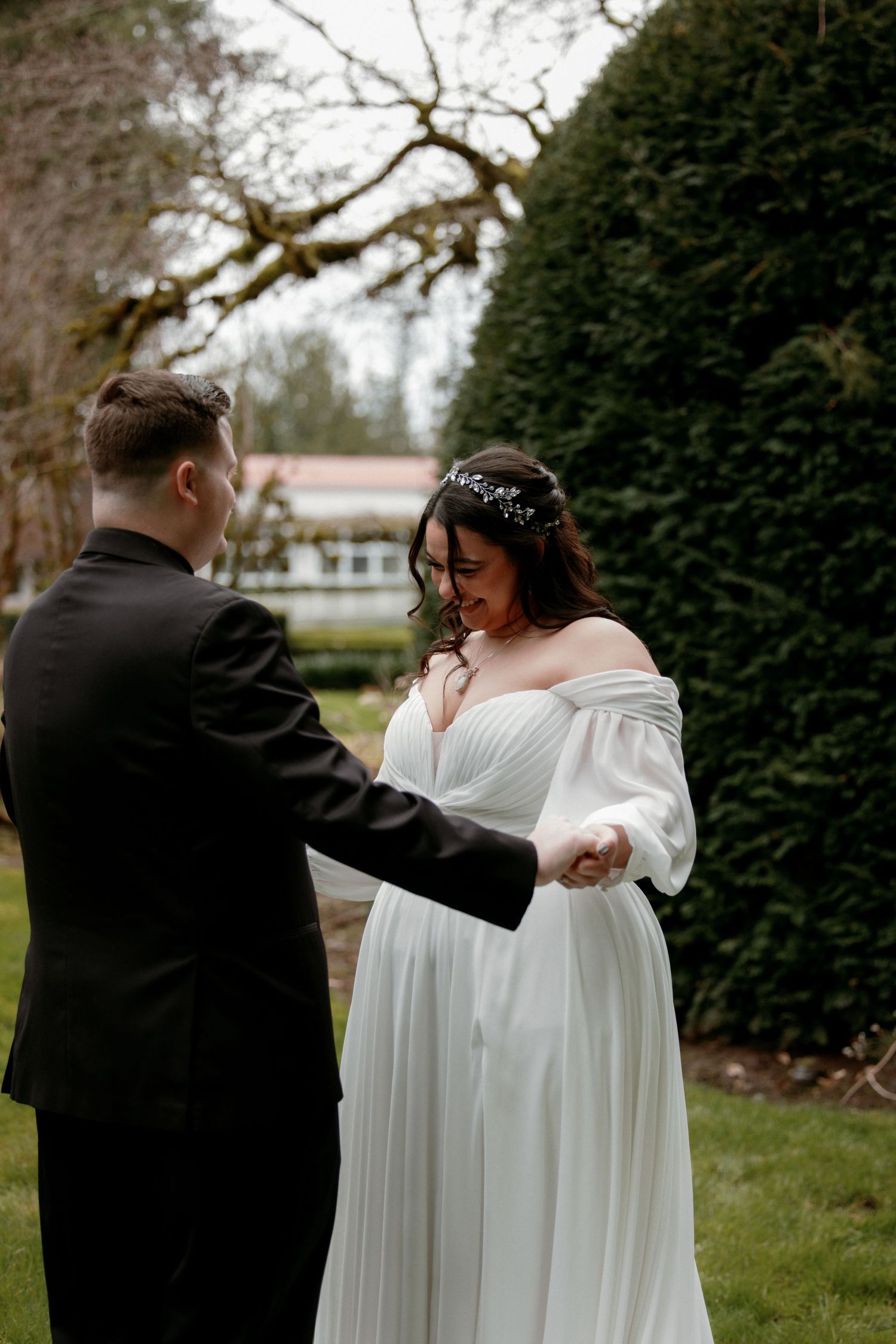 Groom in black suit holding bride's hand as she looks down, standing outdoors among tall hedges.