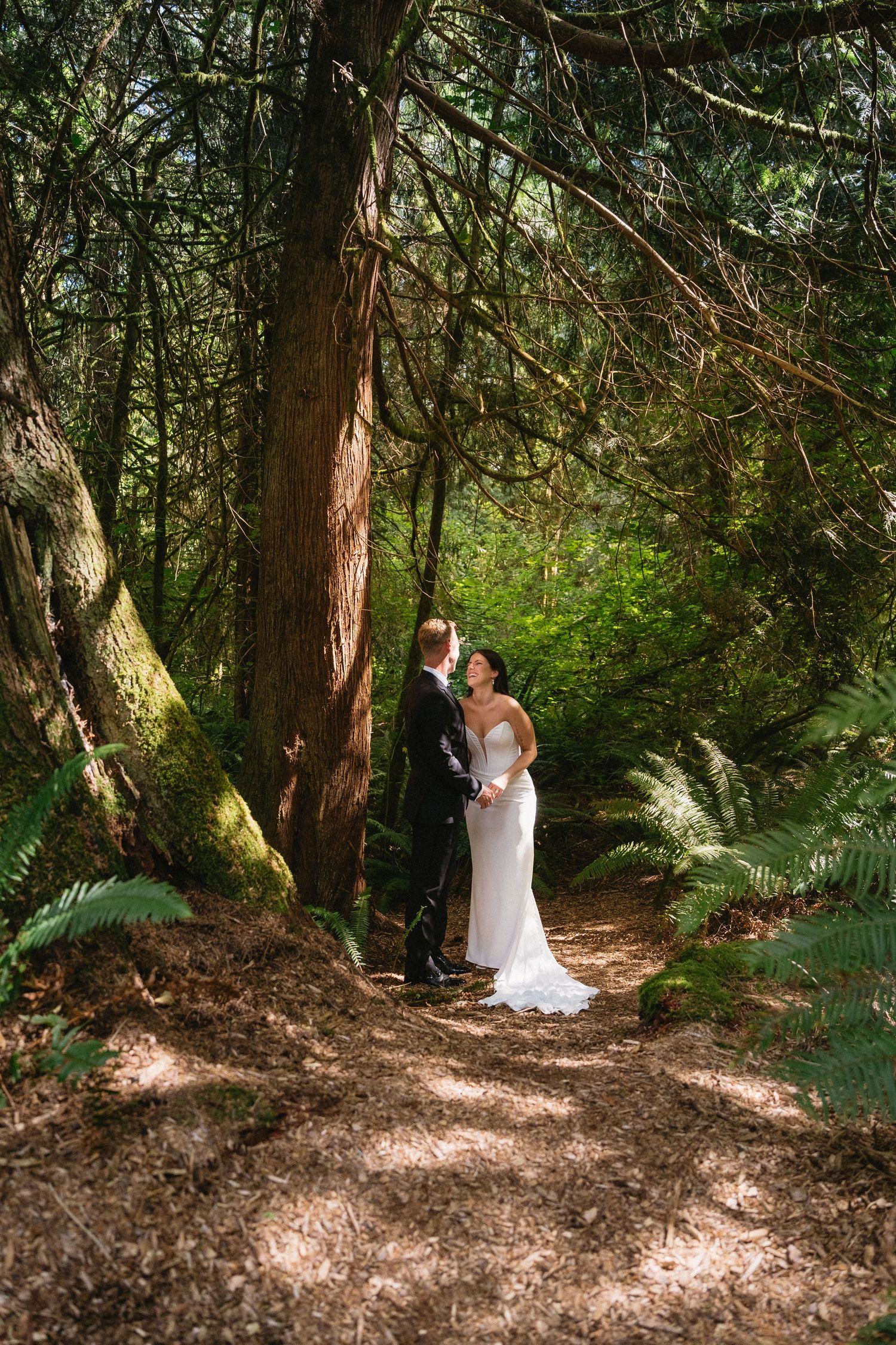 Bride and groom hold hands gazing at each other on forest path.