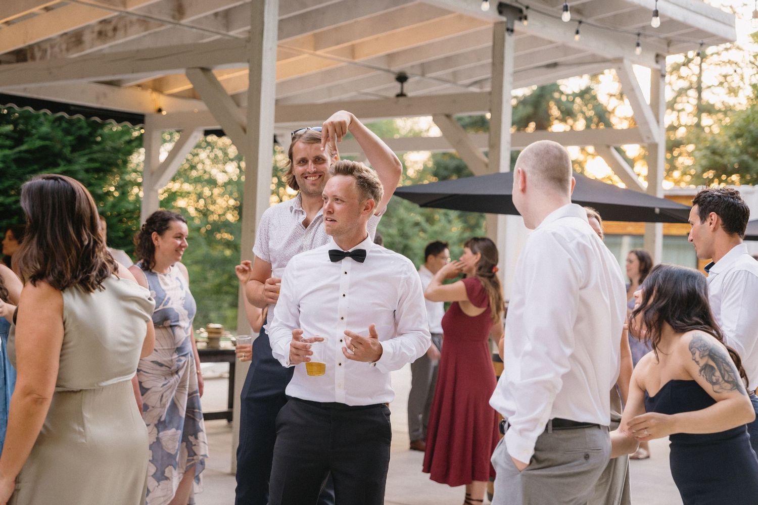 Groom in bow tie laughing with wedding guests during lively outdoor reception celebration.