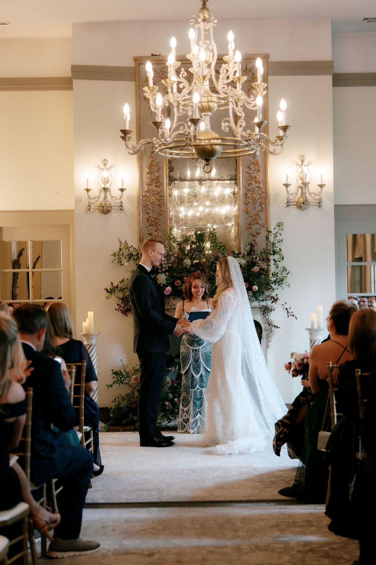 Bride and groom hold hands at the altar beneath a crystal chandelier during an elegant indoor wedding ceremony.