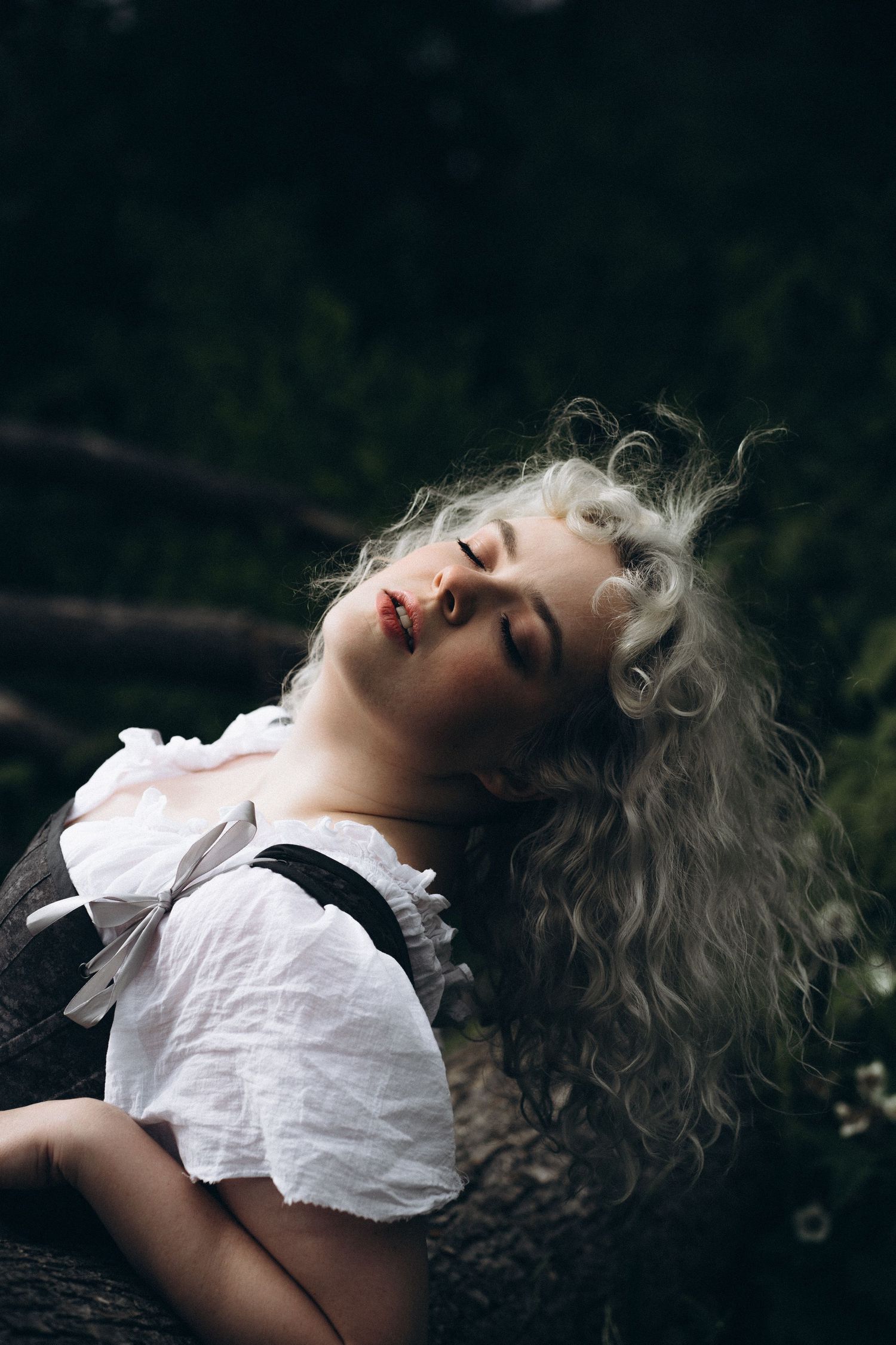 A blonde woman in a white blouse and dark corset rests against a tree trunk with eyes closed in a dark forest.