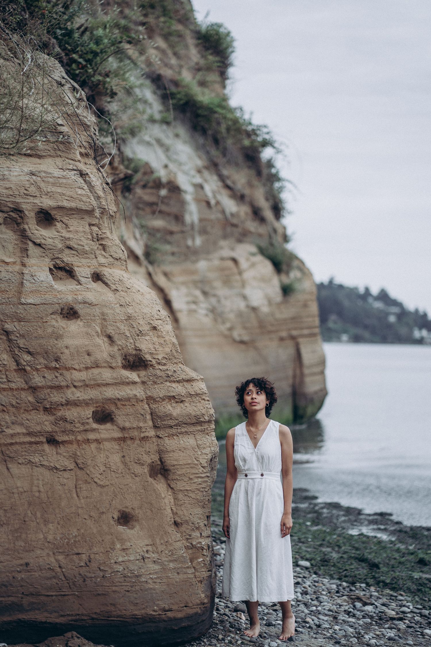 Woman in white belted dress stands barefoot beside tall sandy coastal cliff near water.