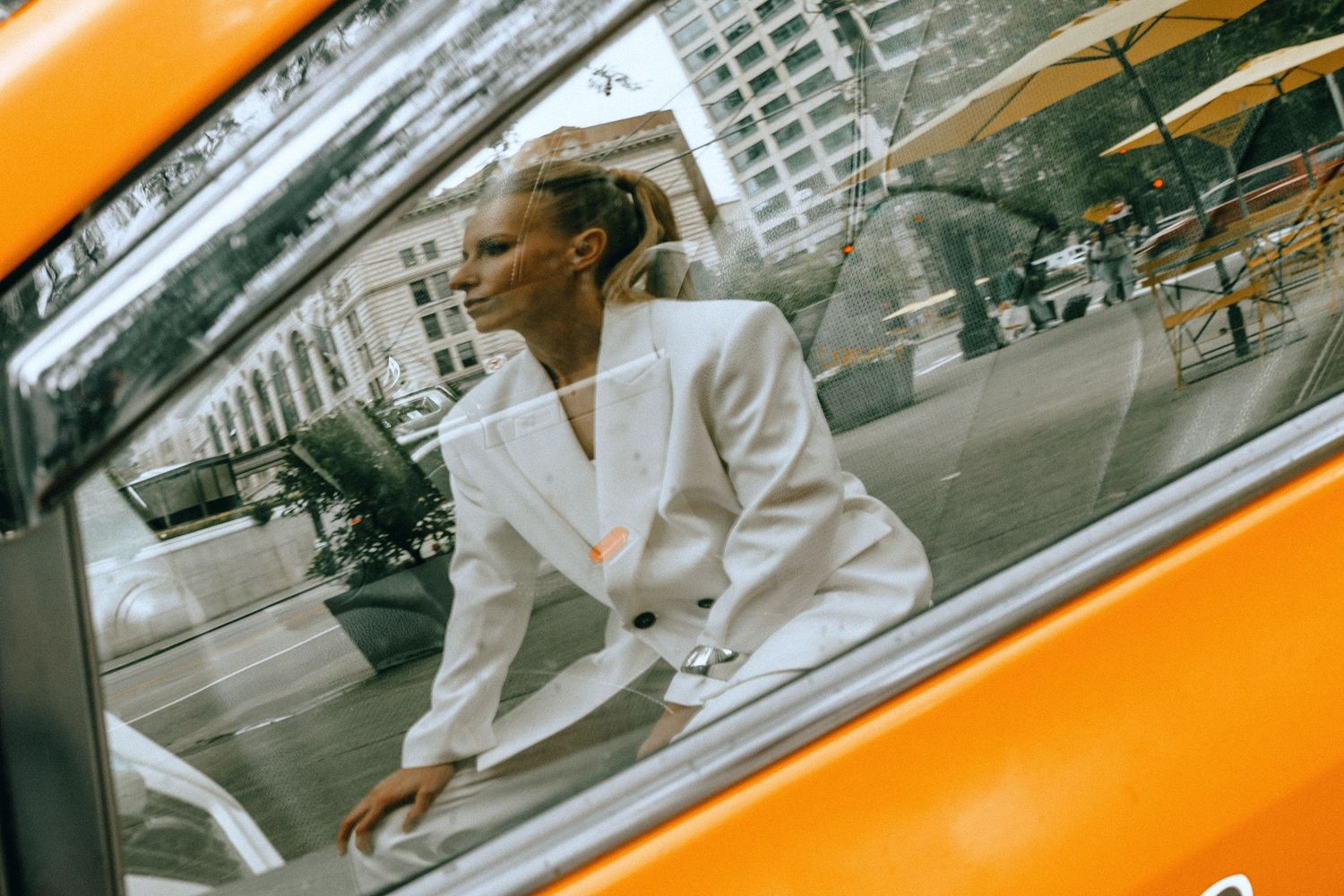 Blonde woman in white blazer photographed through yellow taxi cab window in urban city setting.
