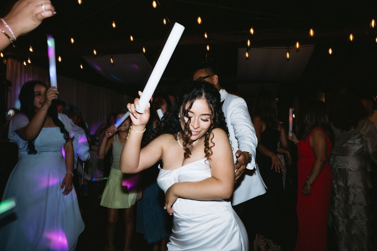 Bride in white gown dances joyfully with a glow stick at a lively wedding reception party.