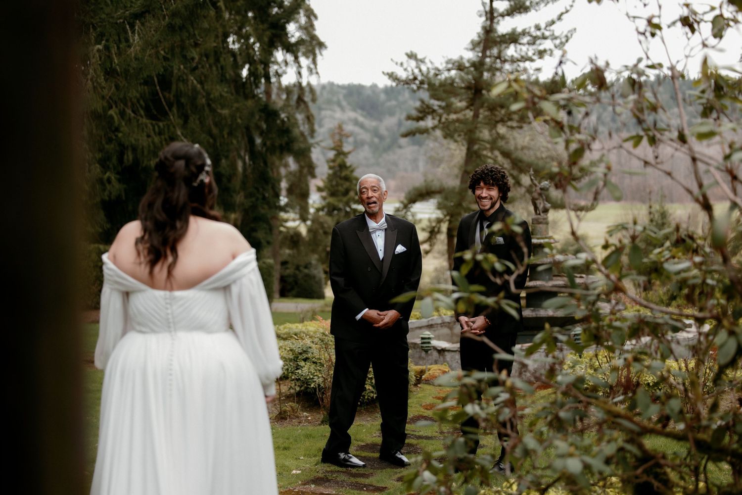 Bride in off-shoulder white gown approaches smiling officiant and witness during an outdoor garden ceremony.