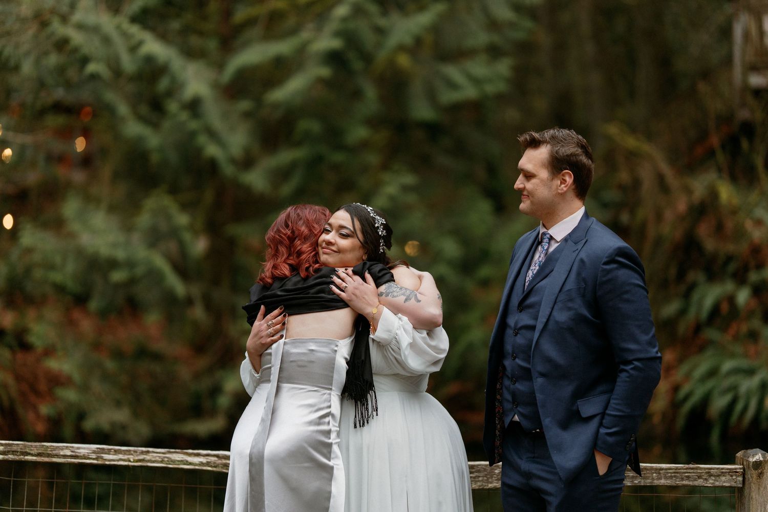 Two brides in white gowns with groom in navy suit at forest wedding.