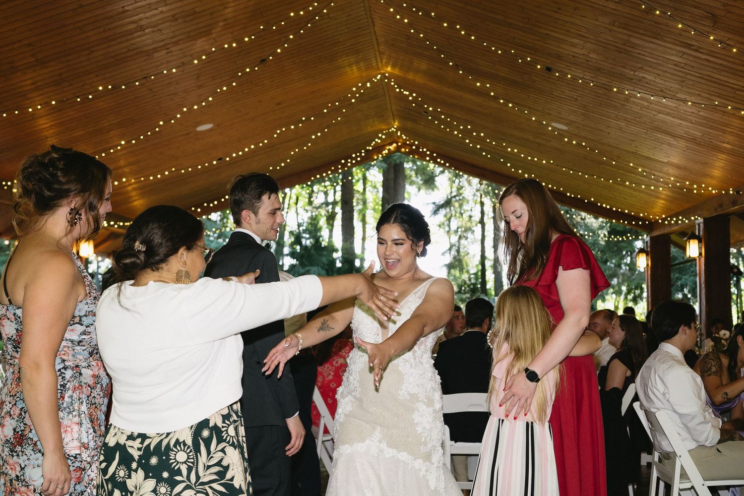 Guests surround and embrace the bride in a lace wedding gown under fairy lights at reception.