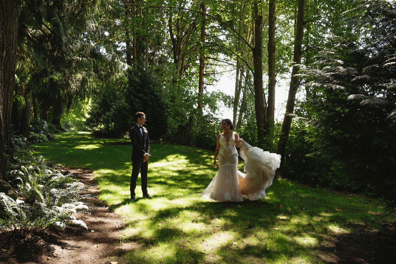 Bride in flowing white gown walks toward groom on a sunlit path through a lush green forest.