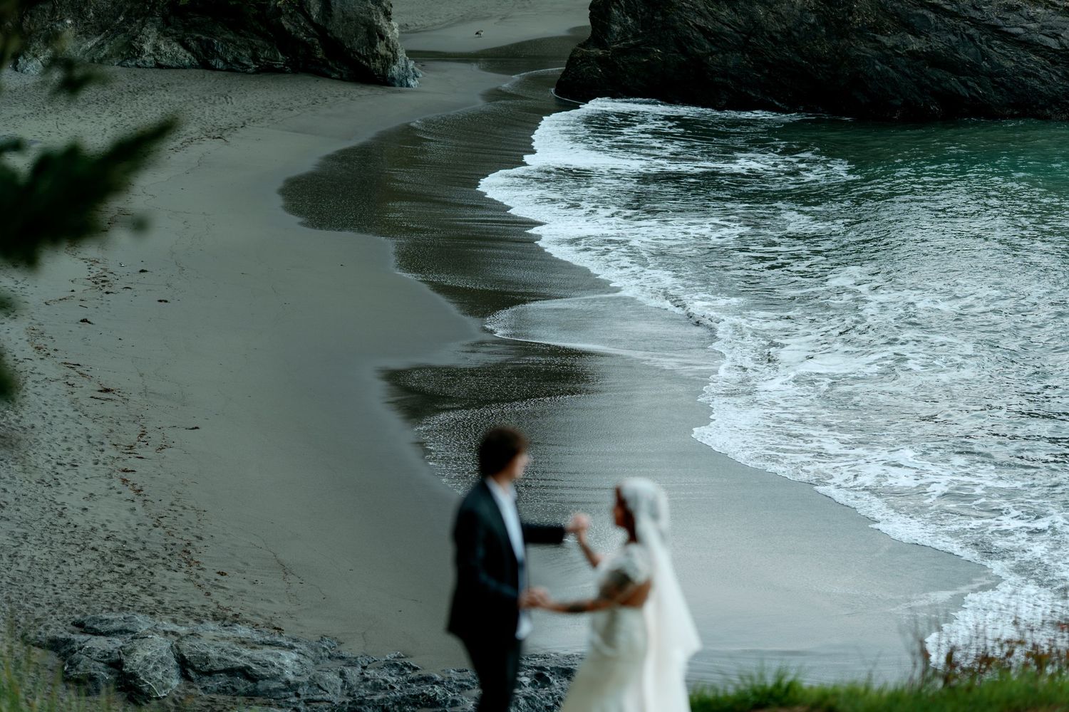 Bride and groom hold hands on a grassy cliff overlooking a serene sandy beach with gentle waves.