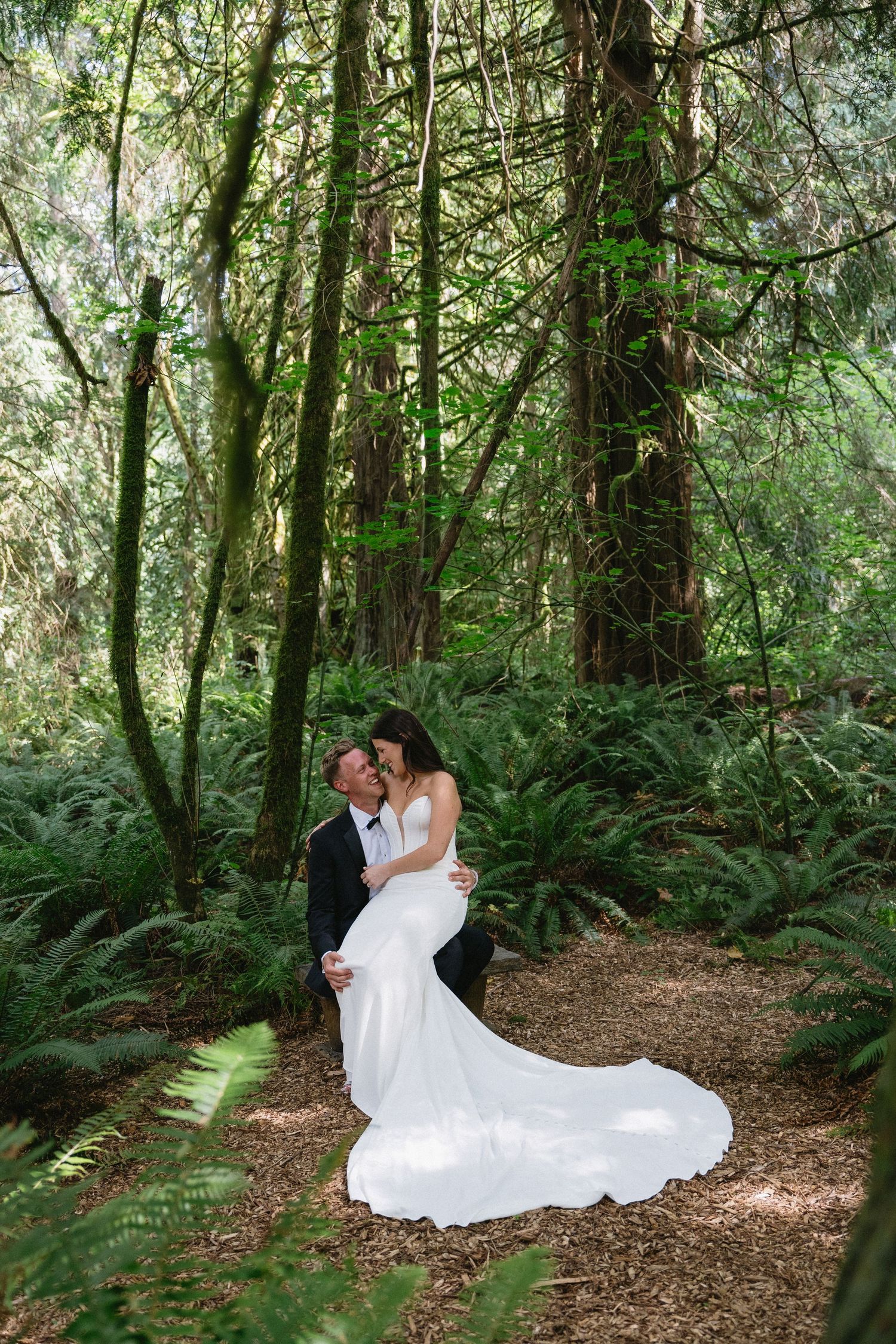 Groom lifts bride in white mermaid gown in lush forest setting.