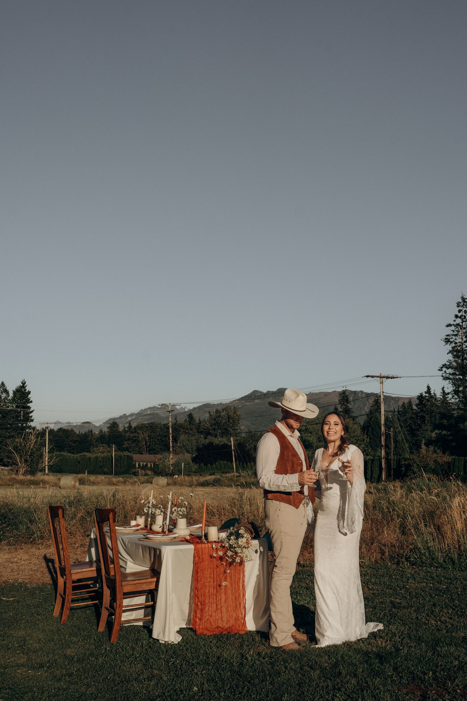 Bride and groom toast at an outdoor rustic tablescape with mountain views at dusk.