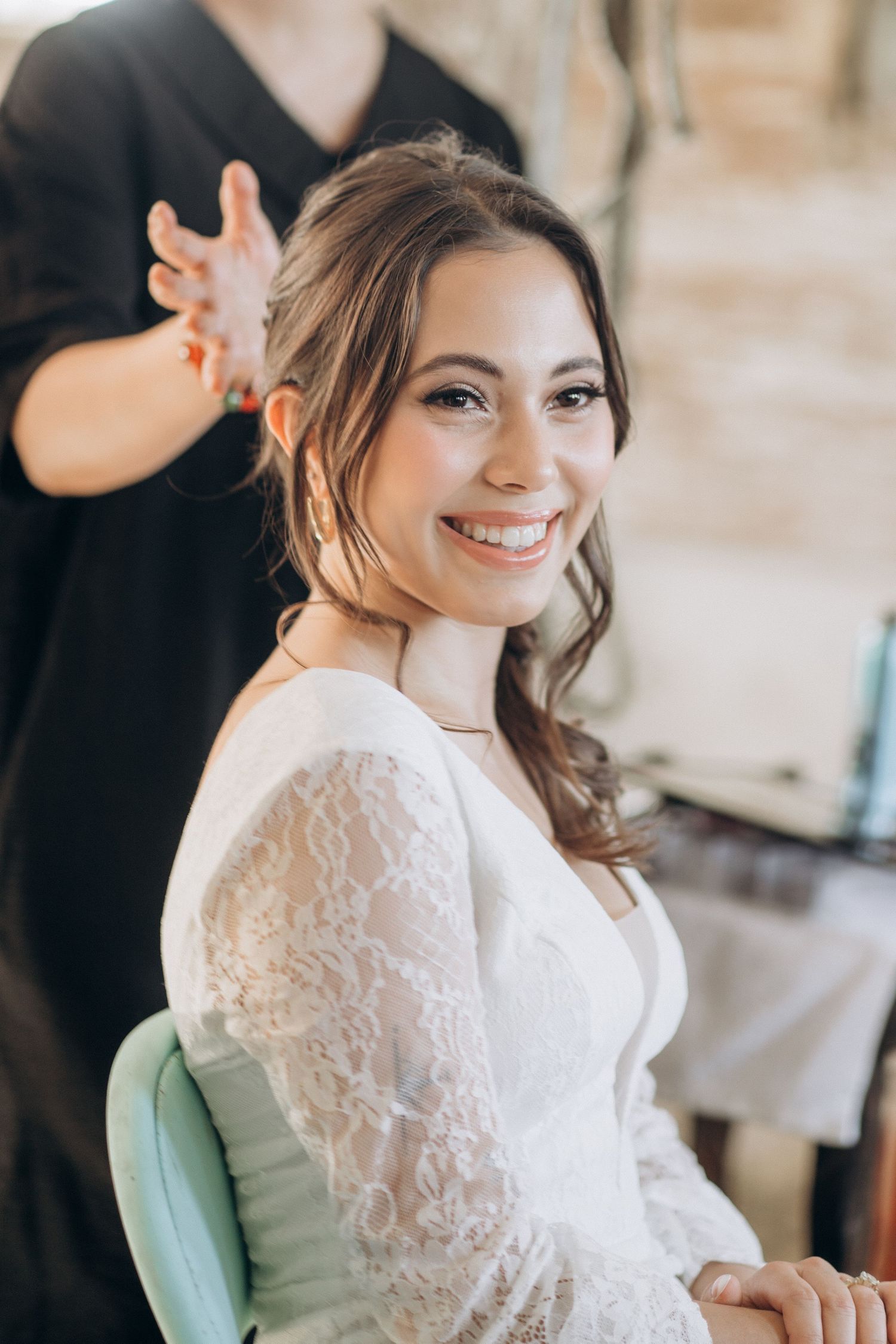 Smiling bride in white lace dress getting hair styled by hairdresser before wedding.