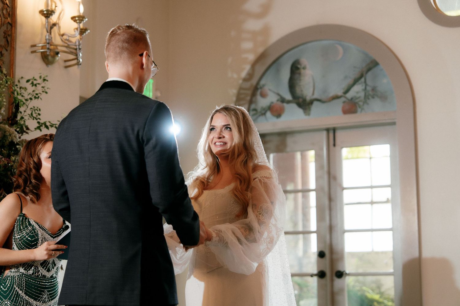 Smiling bride holds grooms hands during indoor wedding ceremony with decorative owl mural visible in background.