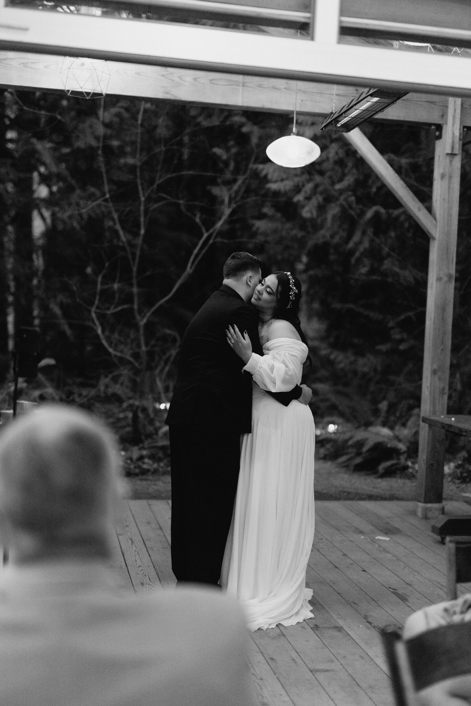 Bride and groom share first dance embrace under wooden pergola at evening wedding reception.