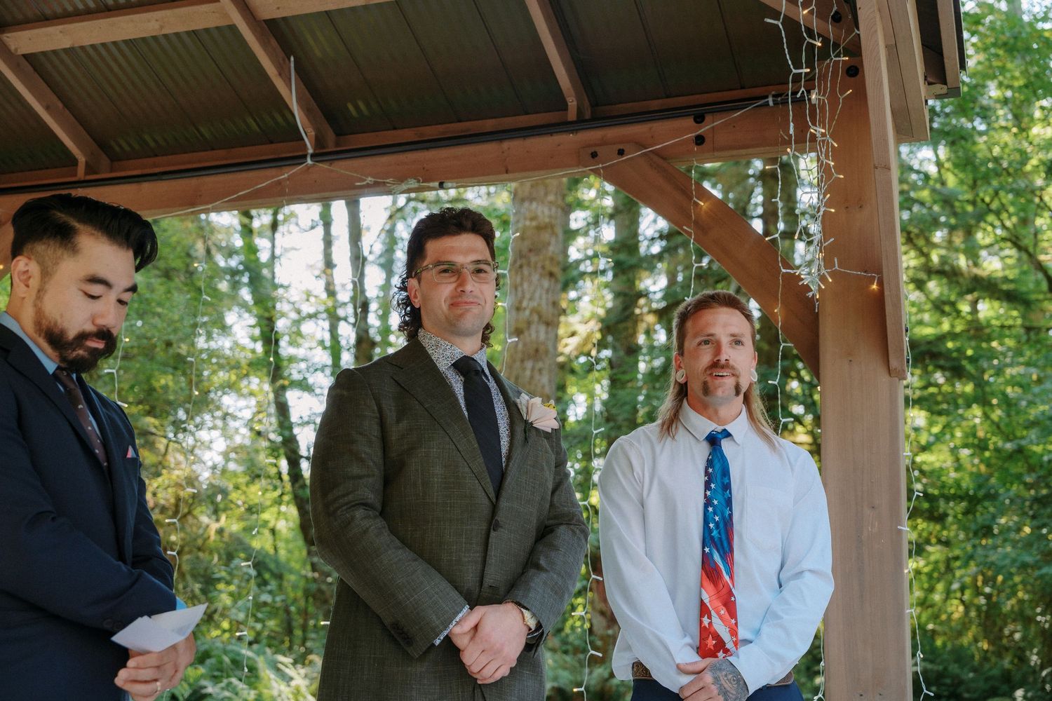 Three men including groom and officiant stand under wooden gazebo at outdoor forest wedding ceremony.