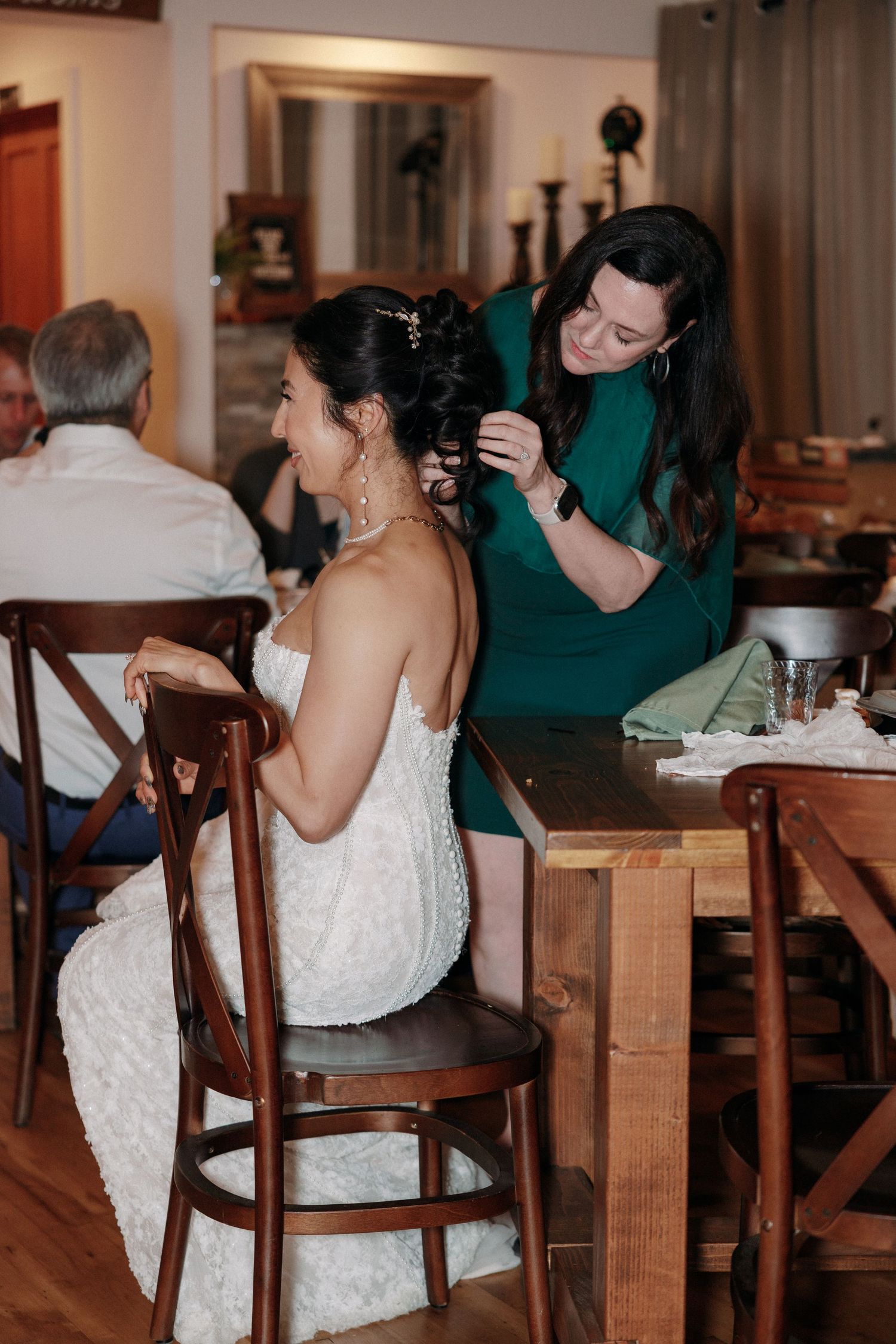 Bridesmaid in green dress adjusting bride's hair at wooden table inside rustic wedding venue.