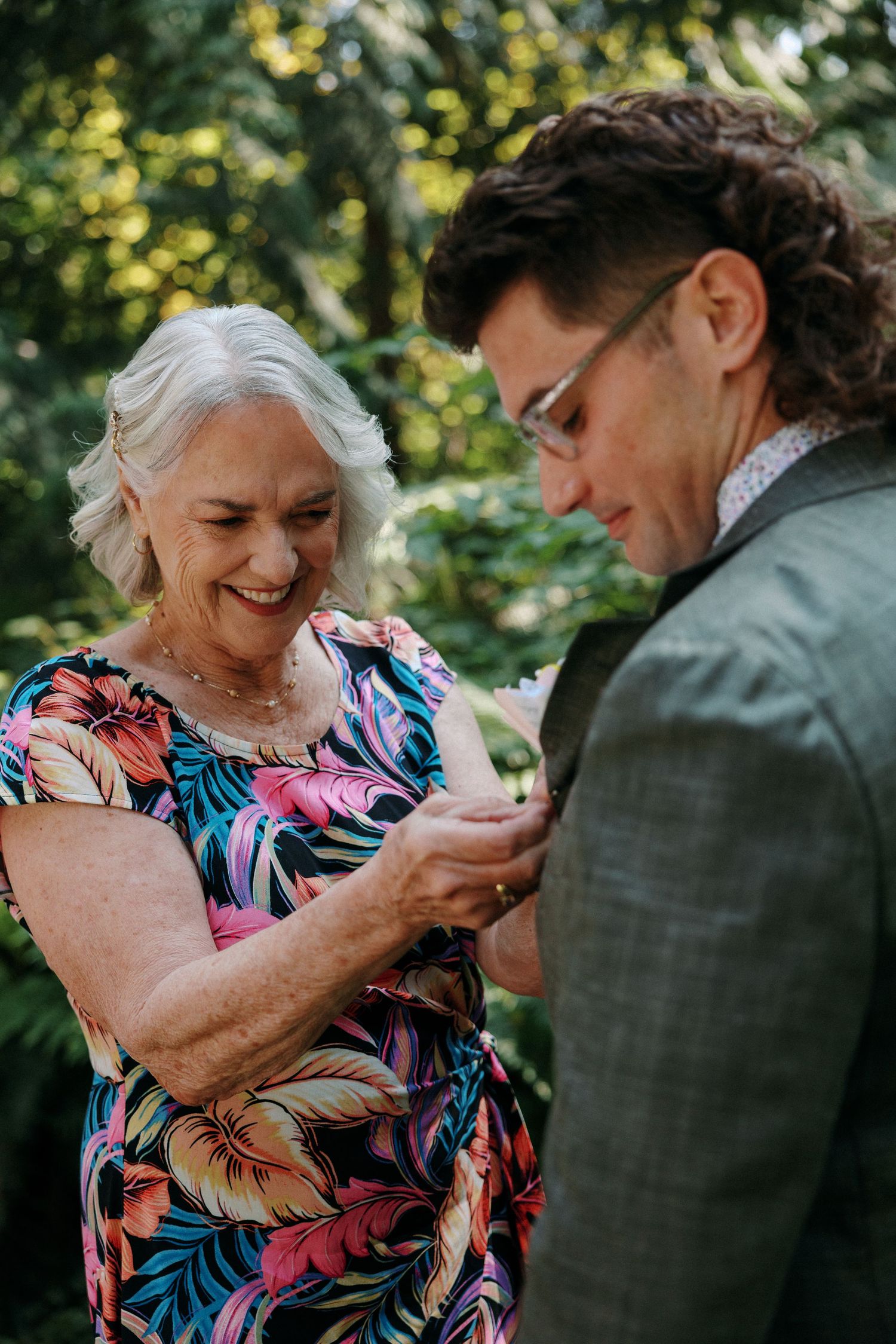 Smiling older woman in colorful floral dress helping groom with boutonniere in lush green garden setting.