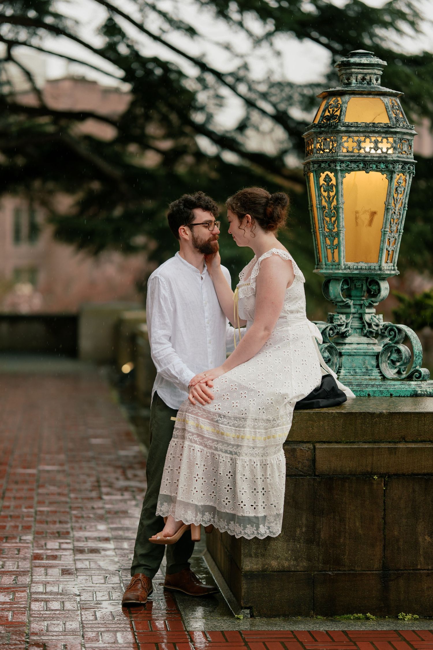 Couple sharing a tender moment beside a glowing ornate lamppost on a rainy day.