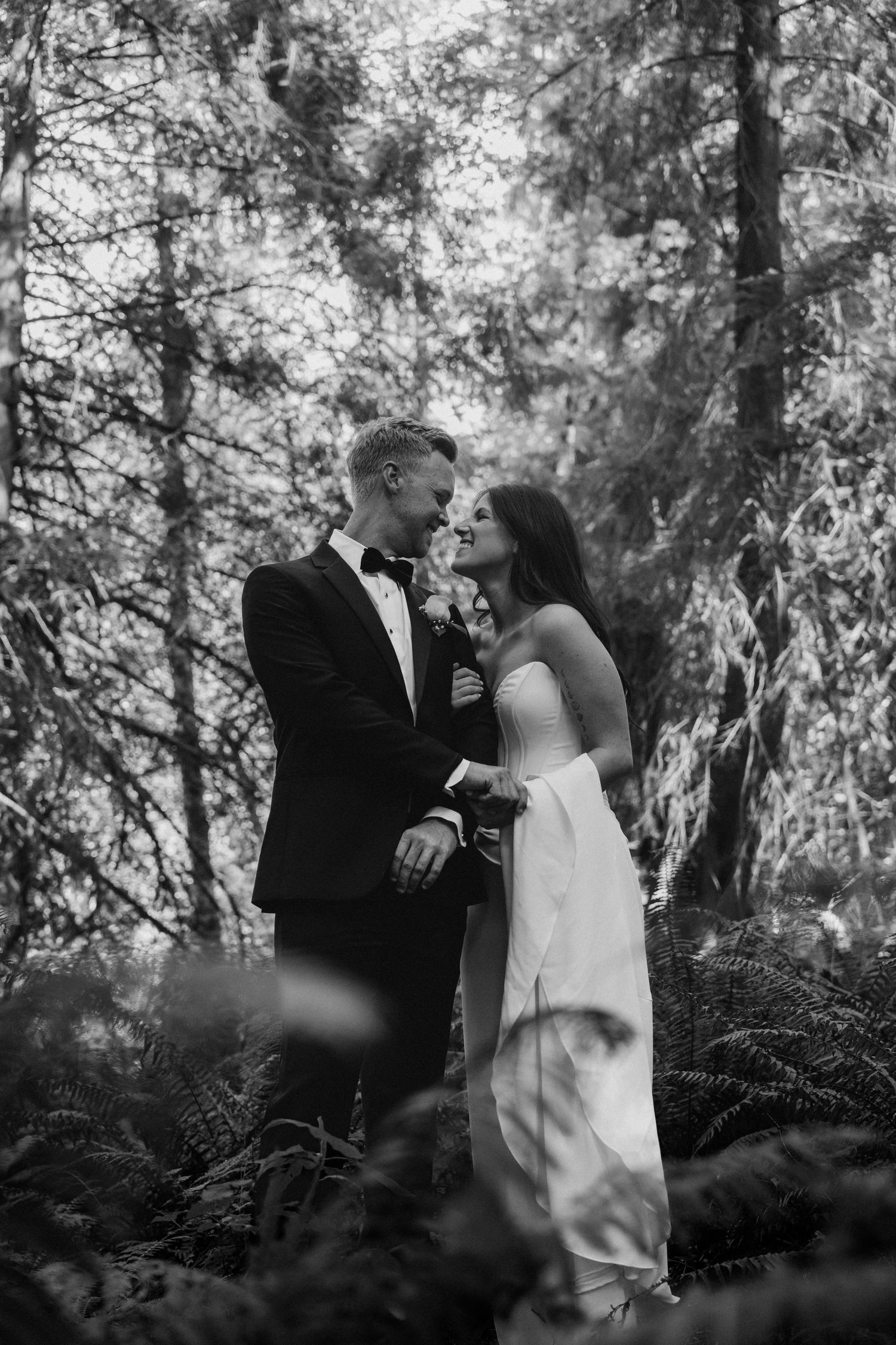Bride and groom share intimate moment surrounded by ferns and tall trees in black and white forest portrait.