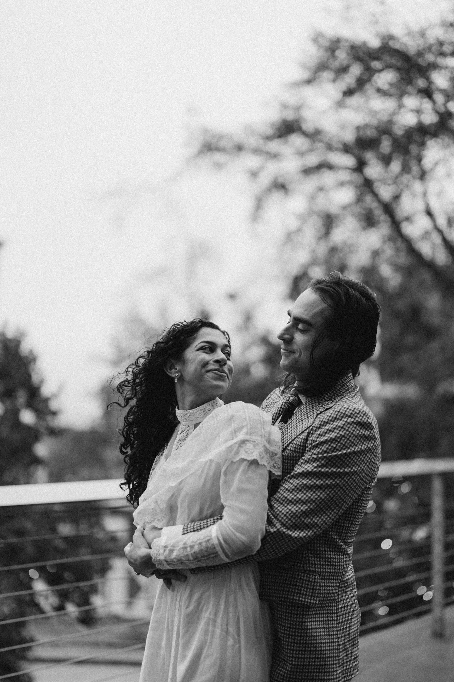 Black and white photo of couple embracing on a bridge, woman in white dress, man in plaid blazer.