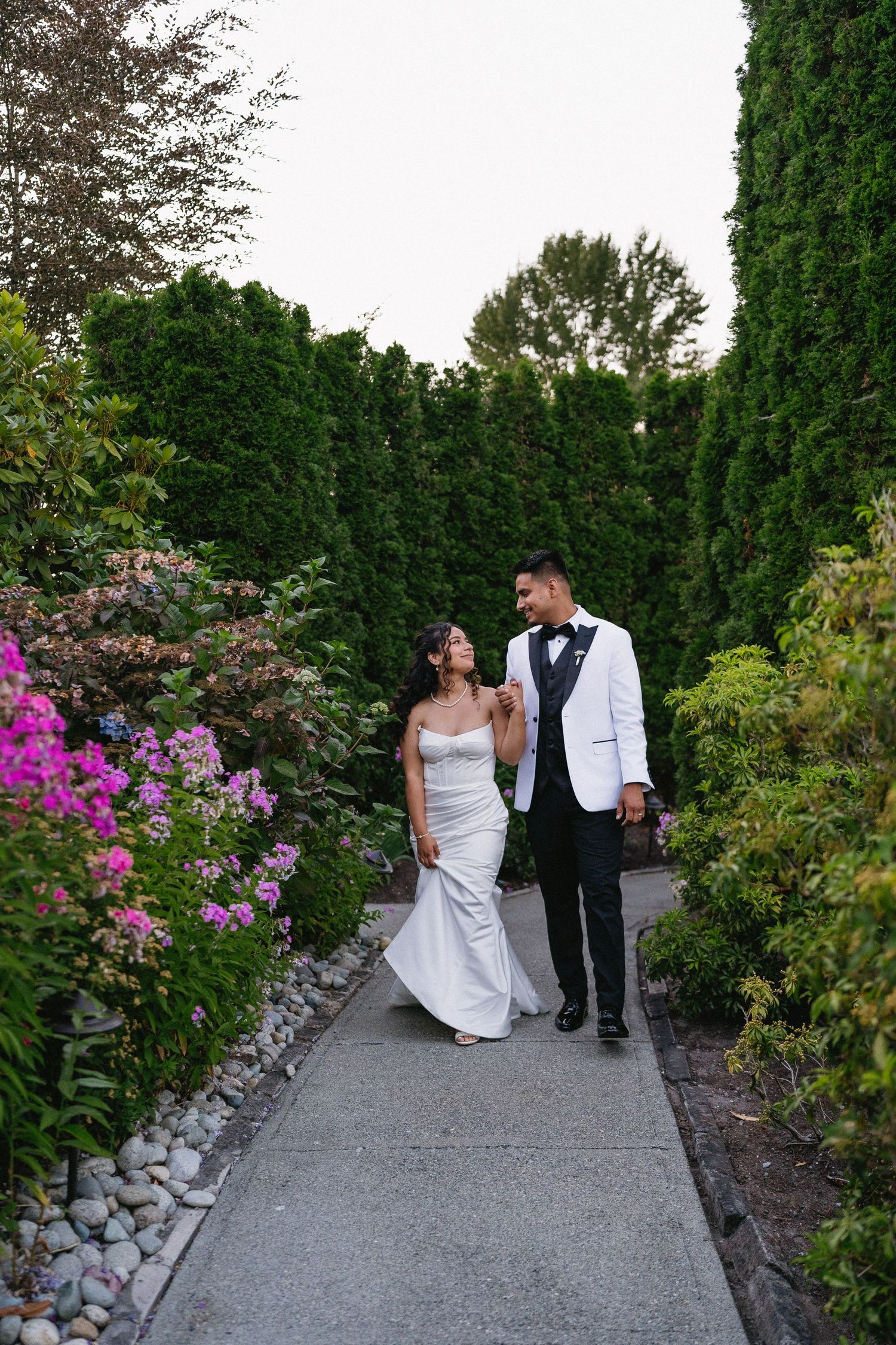 Bride and groom stroll along a lush garden pathway surrounded by tall hedges and colorful flowers.
