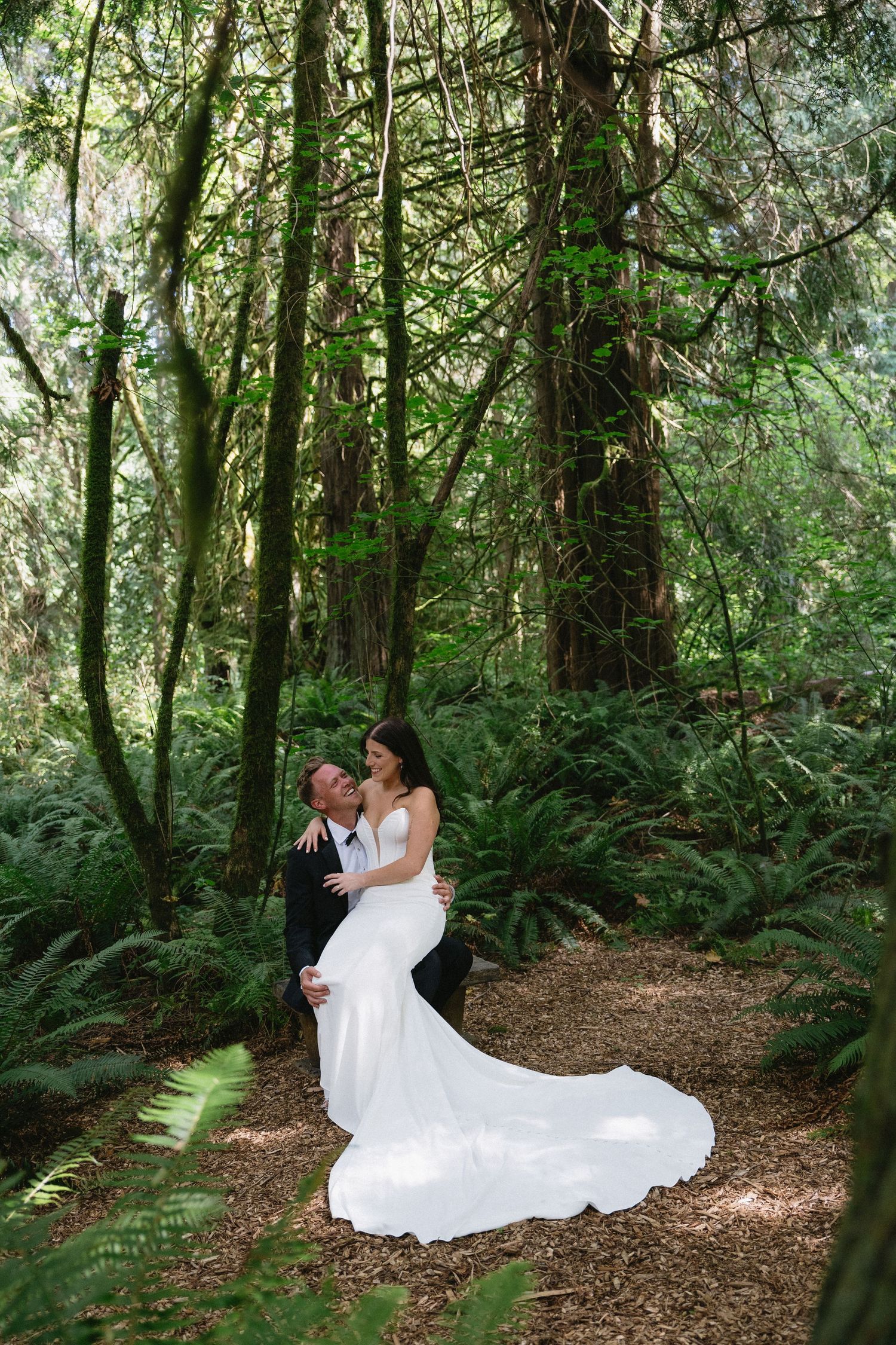 Groom lifting bride in white mermaid gown among lush ferns and towering trees in a forest wedding photo.