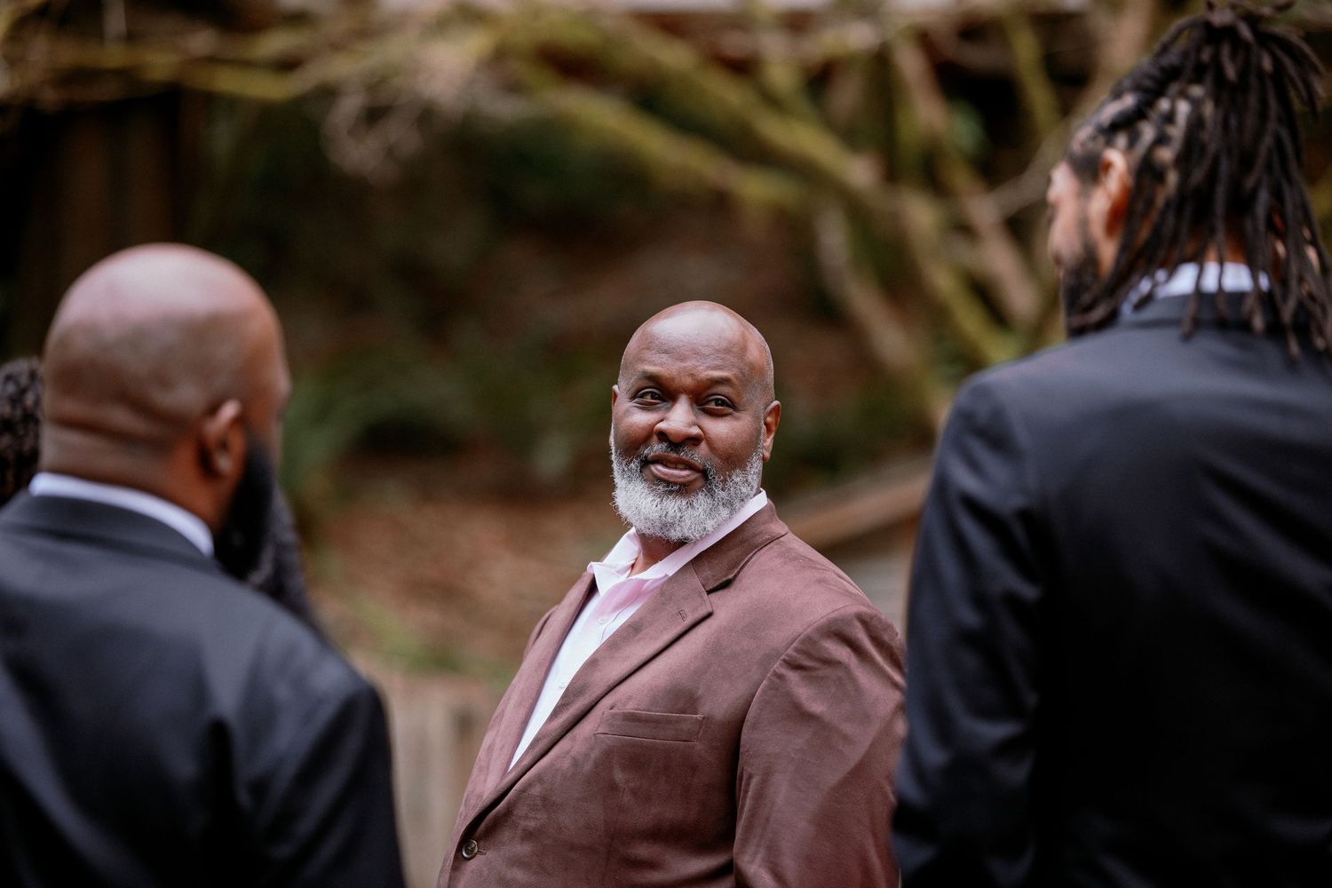 Elegant older man in brown suit smiling warmly at guests during an outdoor wedding.