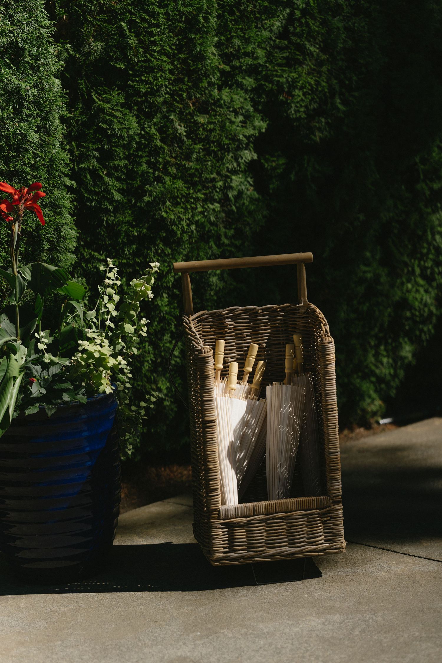 Wicker basket holding wedding programs near lush green hedges at outdoor ceremony entrance.
