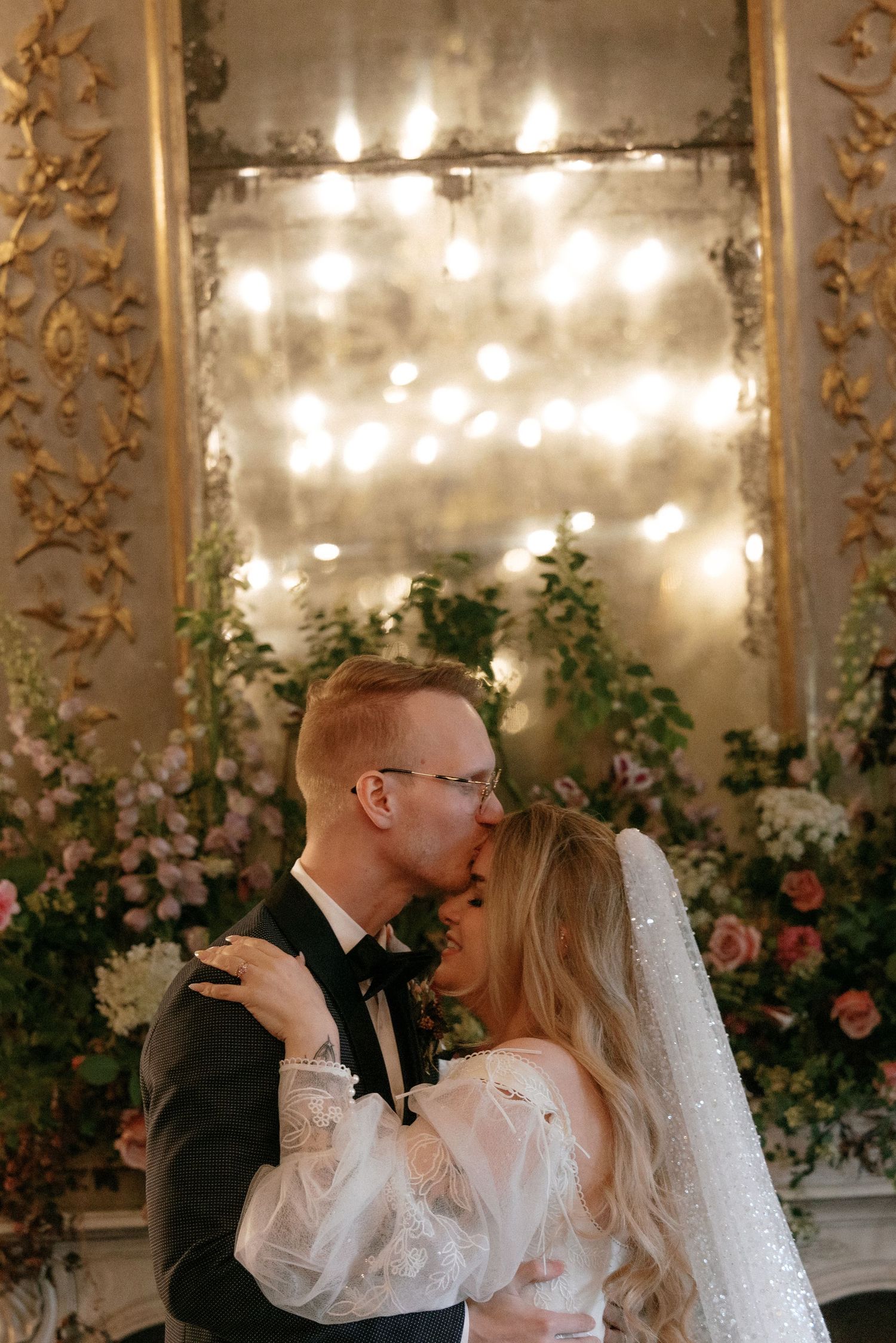 Bride and groom kiss passionately before glowing mirror and pink floral arrangements at wedding.