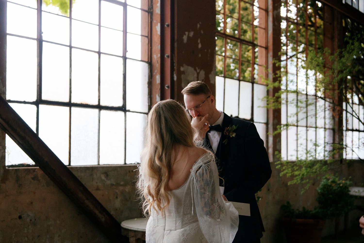 Groom tenderly kisses bride's hand in a rustic industrial wedding venue with large windows.
