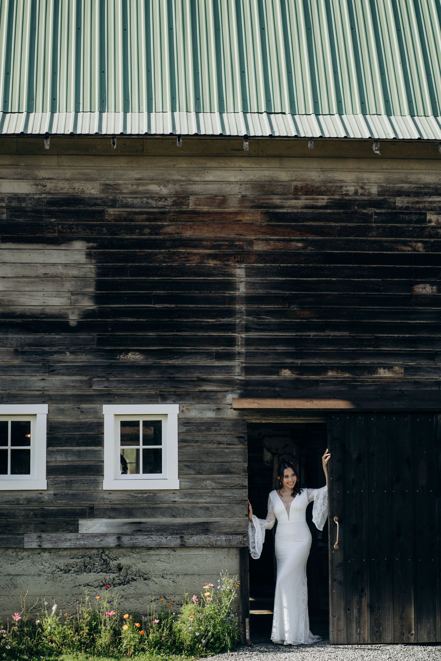 Bride in white lace gown standing in rustic barn doorway surrounded by wildflowers.