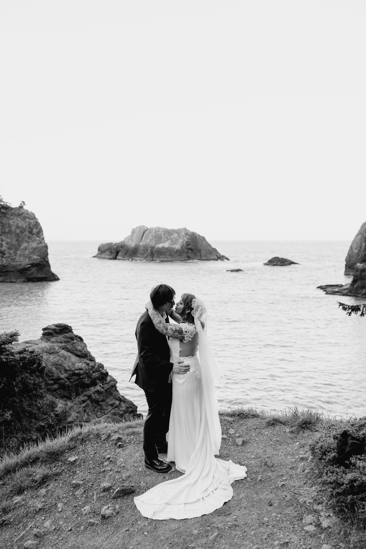 Bride and groom share a romantic kiss on a rugged coastal cliff with ocean rocks behind them.