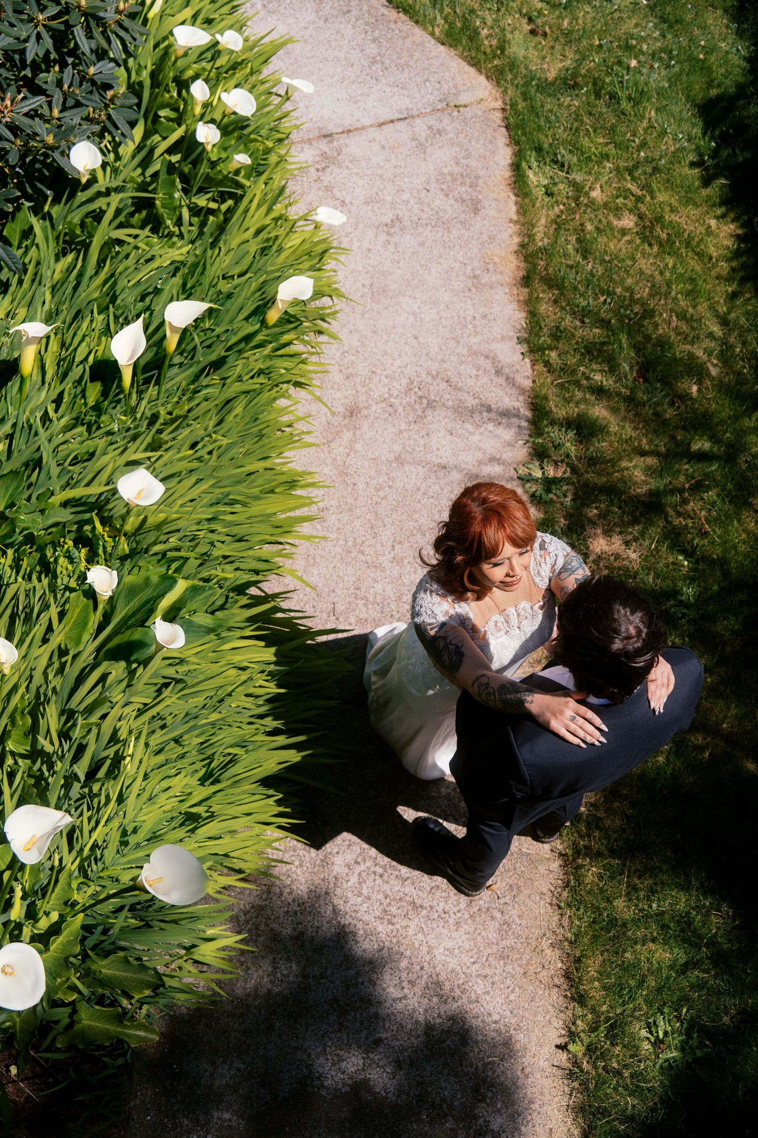 Aerial view of bride and groom embracing on garden path lined with white calla lilies.
