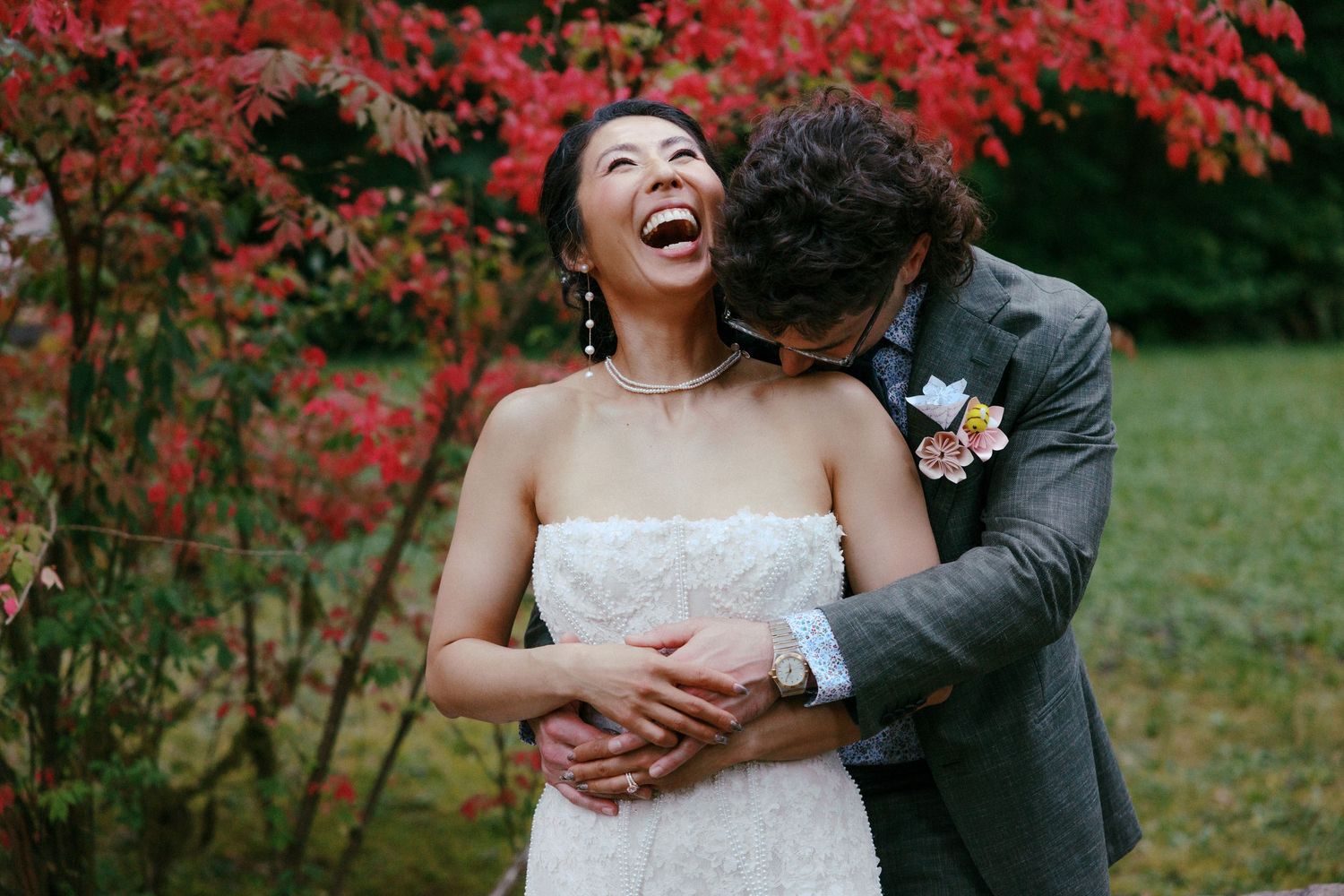 Laughing bride in strapless lace gown embraced by groom near vibrant red autumn foliage outdoors.