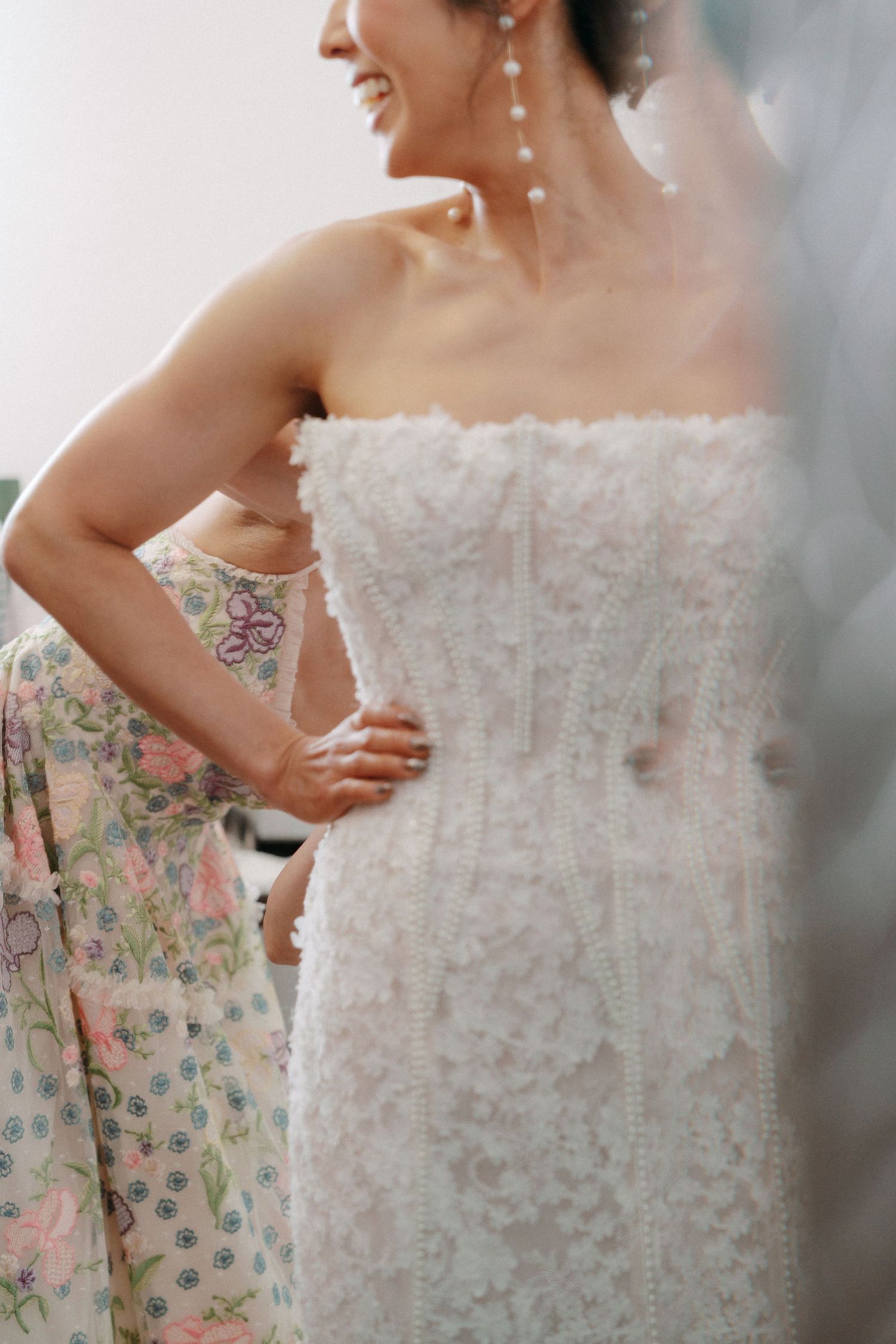 A bride in a strapless white lace wedding gown laughs while being helped by a woman in a floral dress.