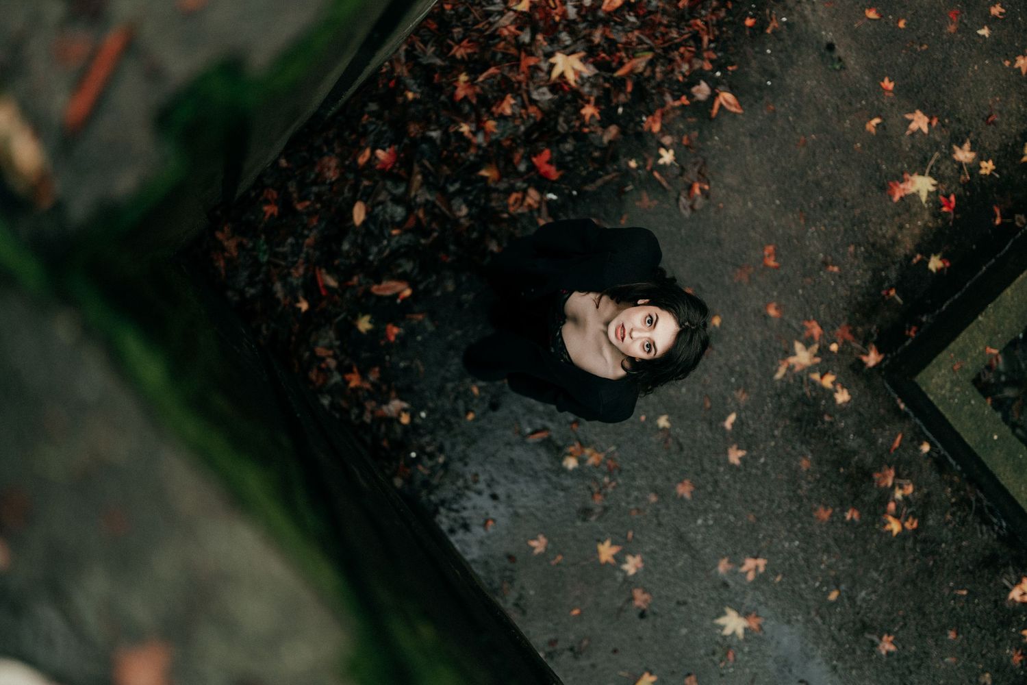 Aerial view of woman in black outfit standing on wet pavement surrounded by fallen autumn leaves.