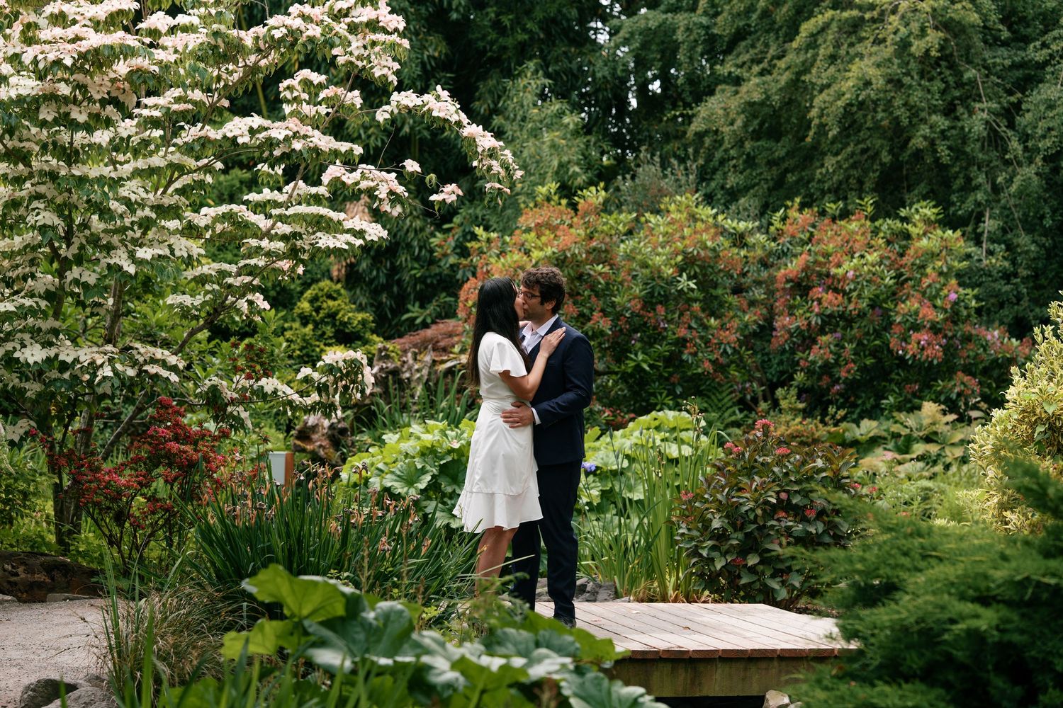 Couple sharing a kiss in a lush garden with white flowering shrubs and colorful plants surrounding them.