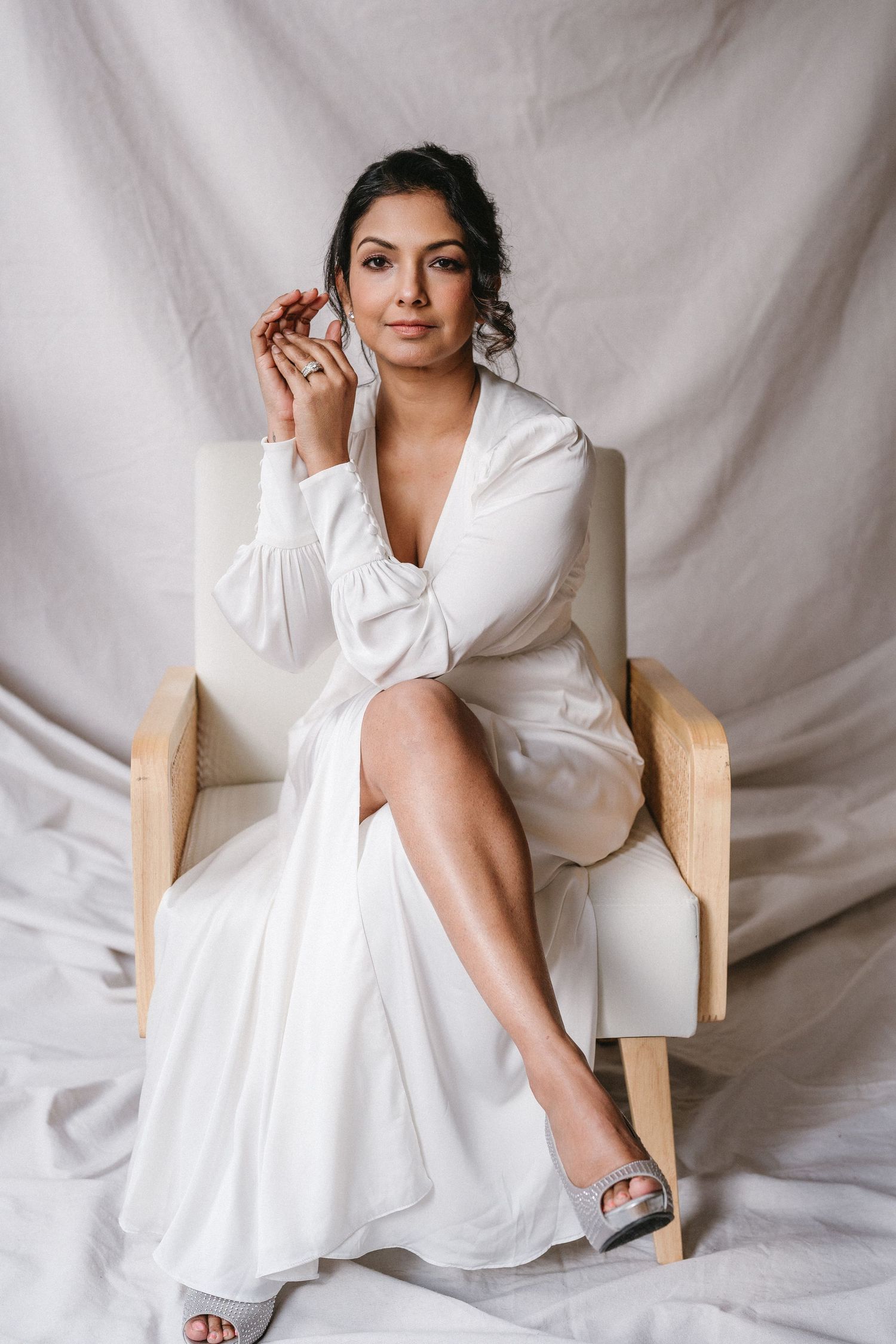 Bride in white satin wrap dress sits in armchair holding accessory against draped fabric backdrop.