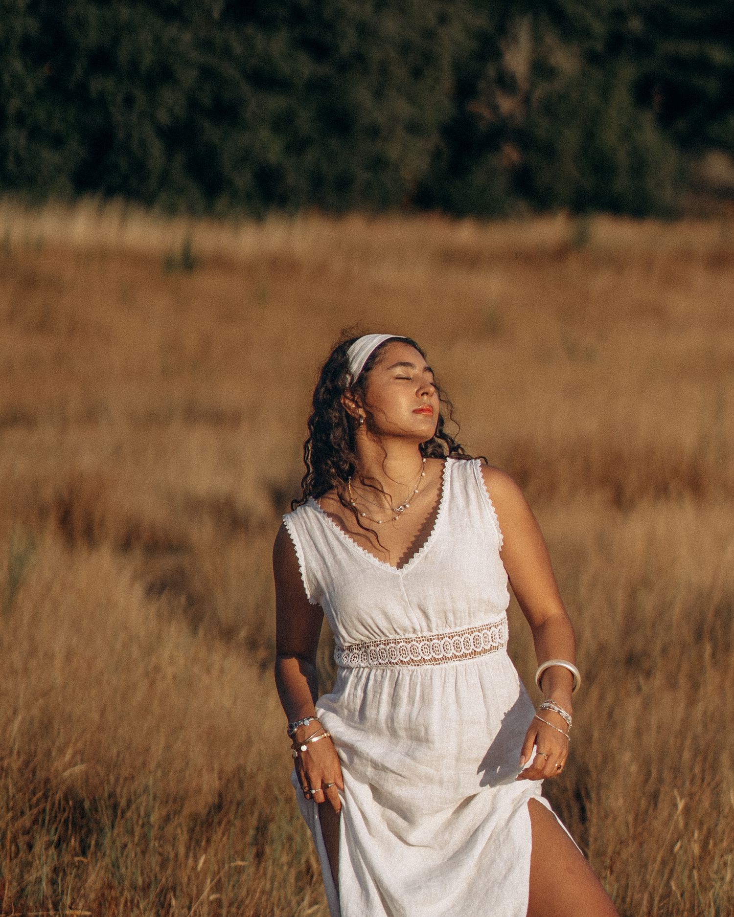 Woman in white maxi dress with headband standing in golden field, eyes closed, enjoying warm sunlight.