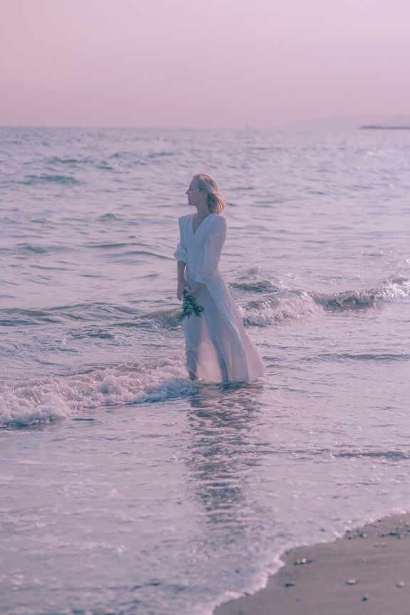 Woman in white dress standing in ocean waves at pink sunset holding flowers.