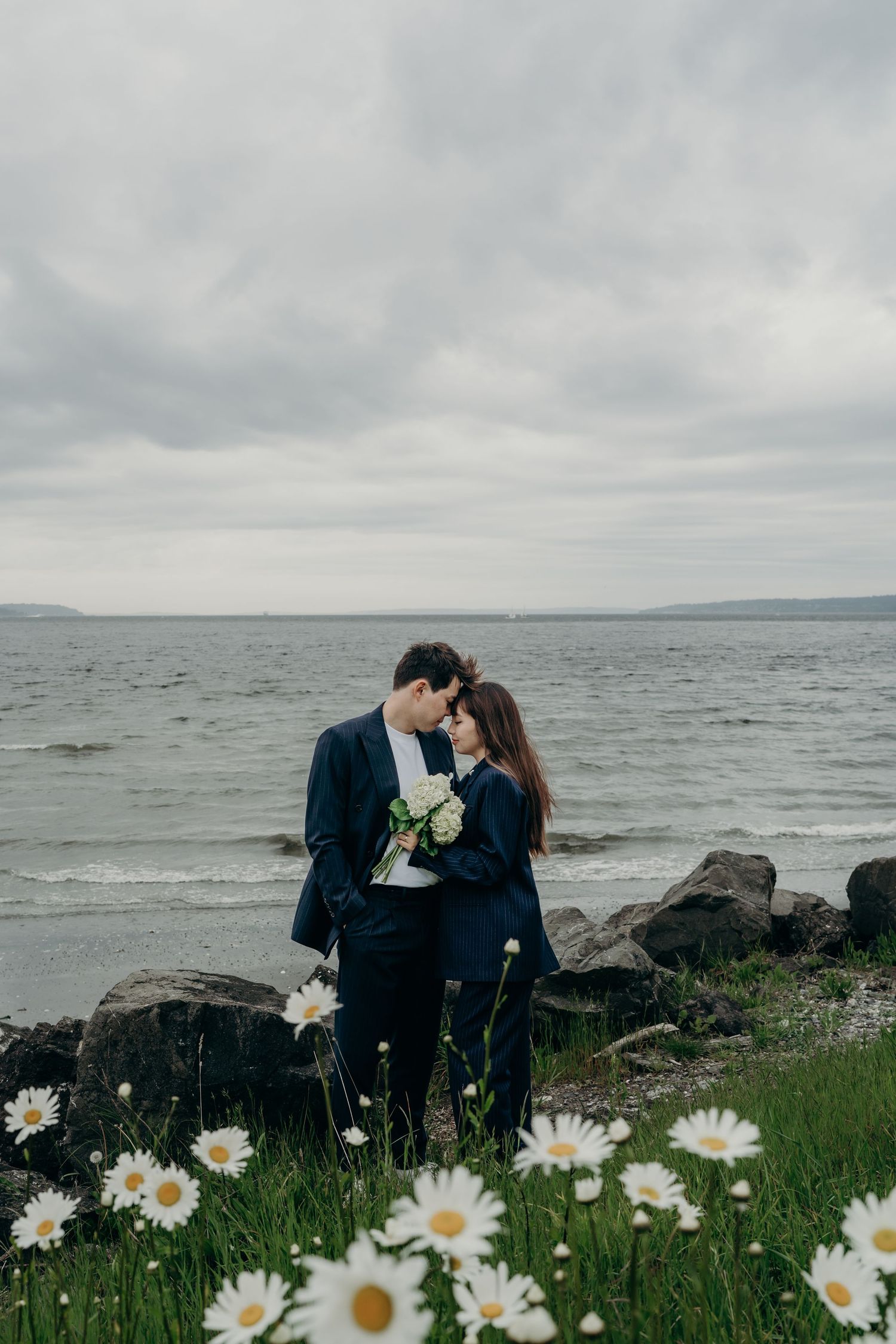 Couple in navy suits sharing intimate moment among white daisies with overcast ocean backdrop.