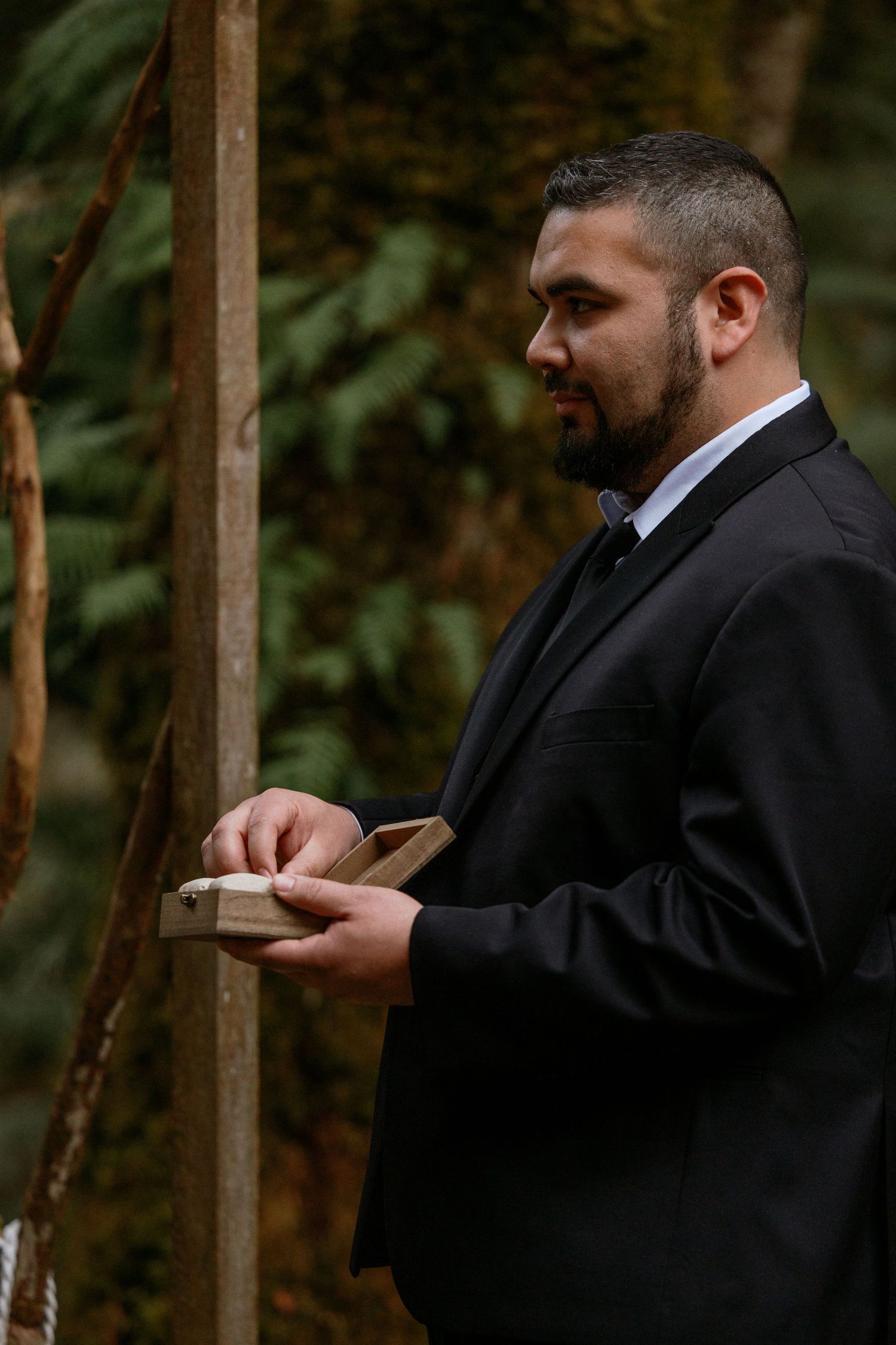 Groom in black suit holding wooden ring box at rustic outdoor forest wedding ceremony.